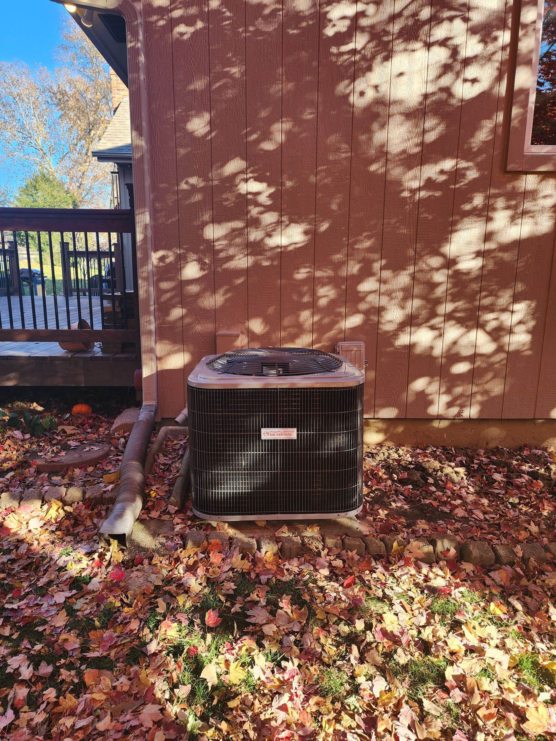 A square outdoor air conditioning unit sits on the ground next to a brown stucco wall, surrounded by fallen autumn leaves.
