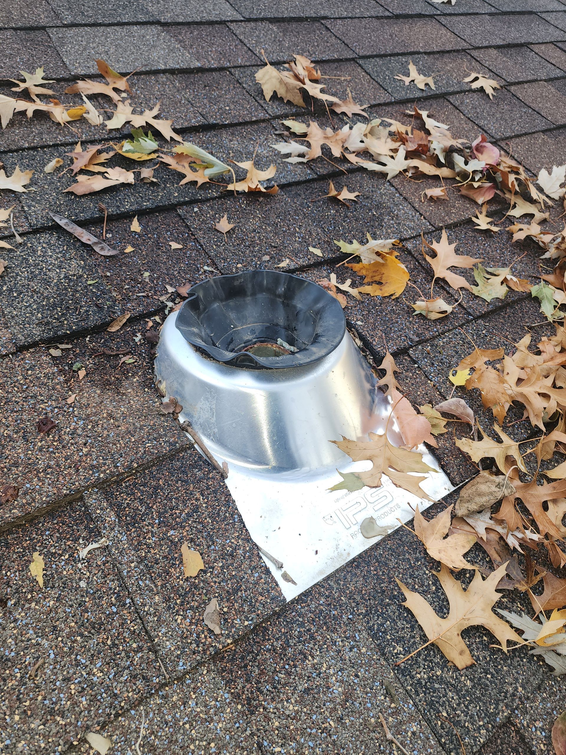A metal roof vent with a black rubber boot sits on asphalt shingles scattered with dried autumn leaves.