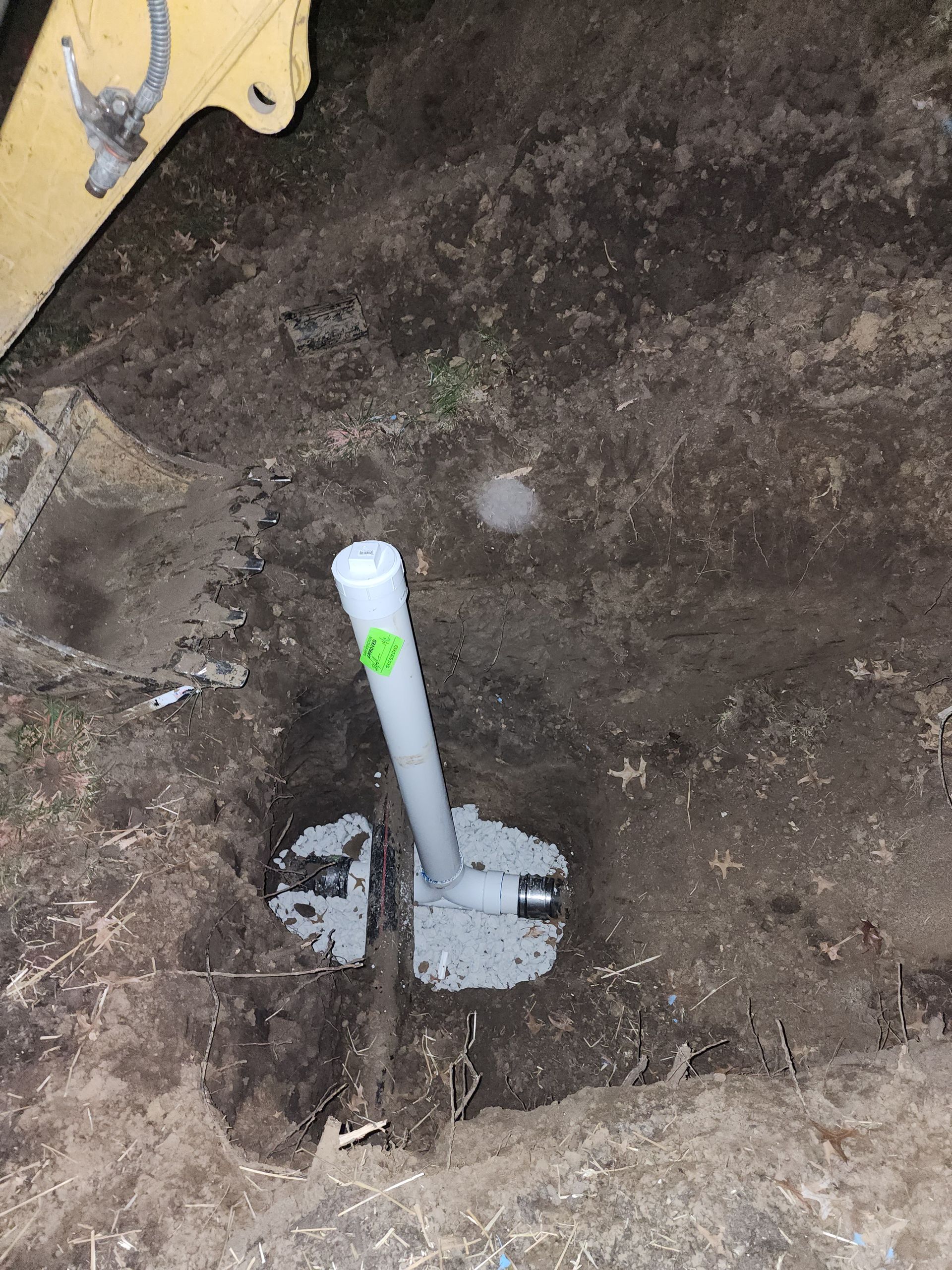 A vertical white PVC pipe stands in a dirt excavation, secured at the base with fresh, light-colored concrete.