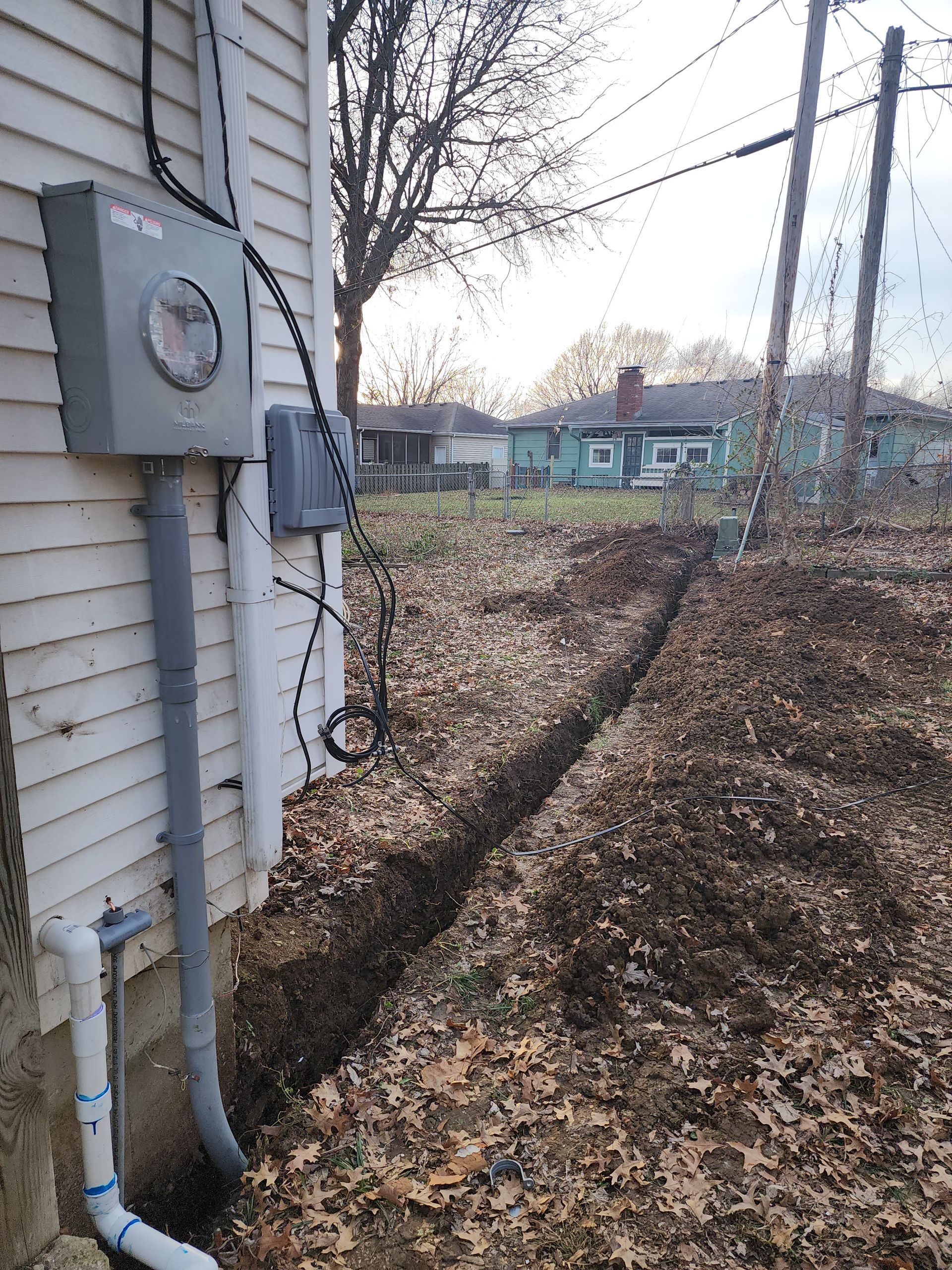 An electrical meter box on a house exterior with a trench dug in the ground leading toward utility poles.