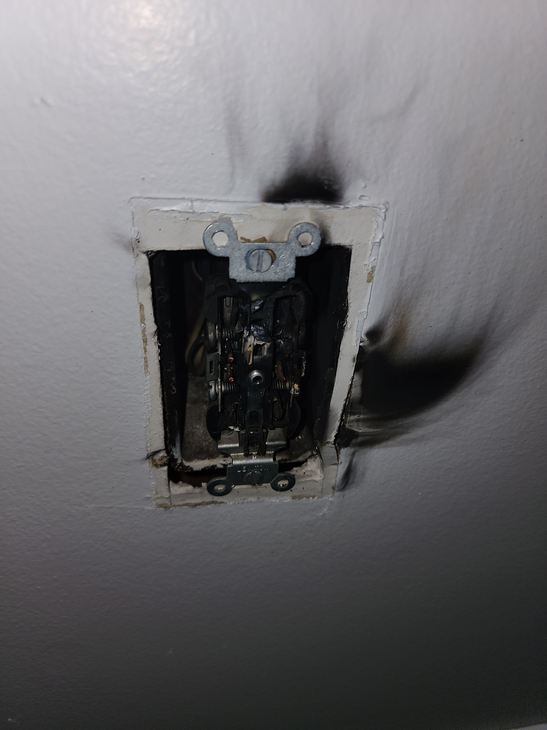 A wall outlet with severe charring and black soot stains surrounding it on a white wall.