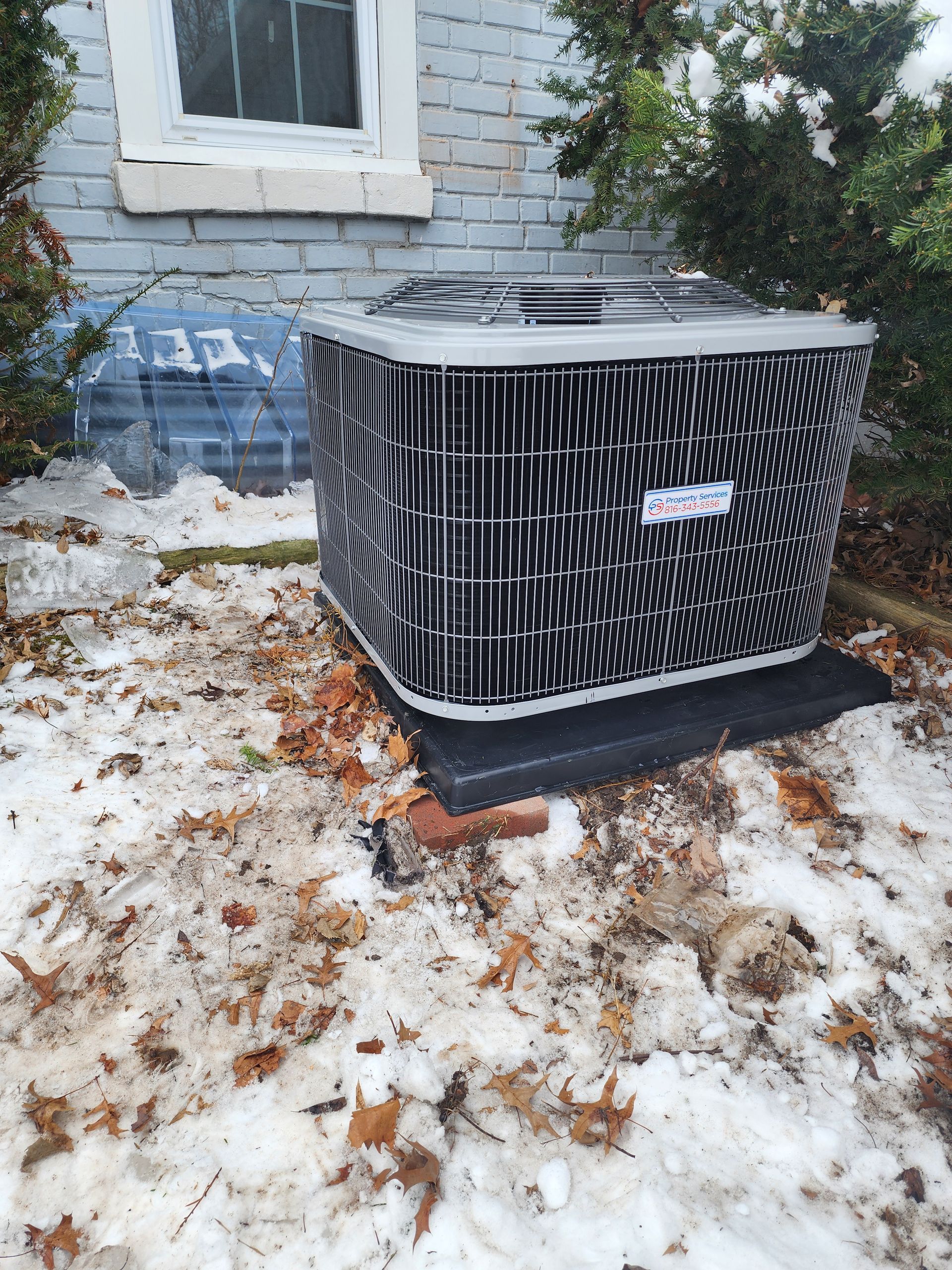 An outdoor HVAC unit sitting on a black pad in a snowy, leaf-covered yard beside a brick house.