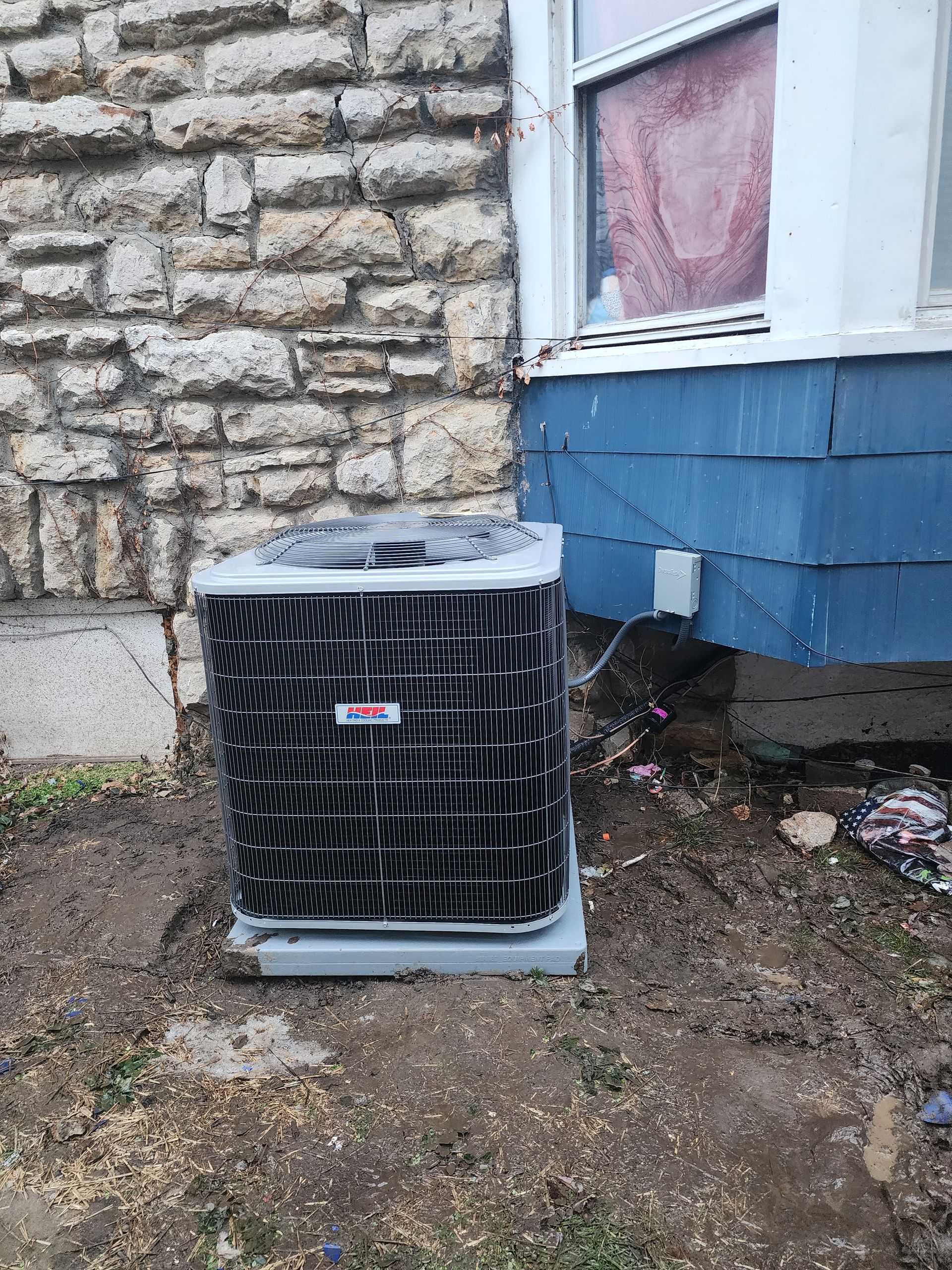 An air conditioning condenser unit sits on the ground next to a stone wall and a blue-sided house.