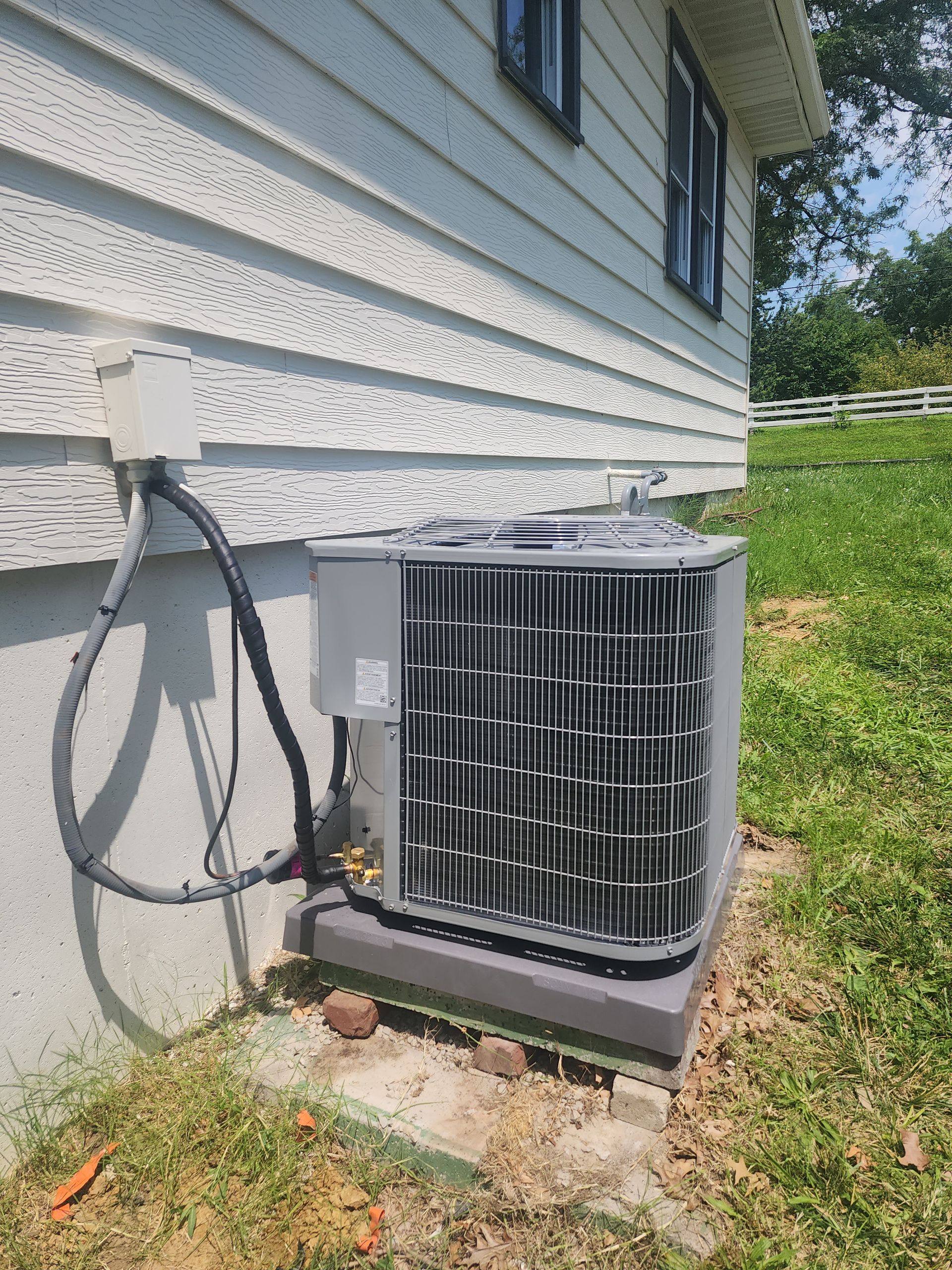 A grey air conditioner unit sits outside a white-sided house on a concrete slab foundation.