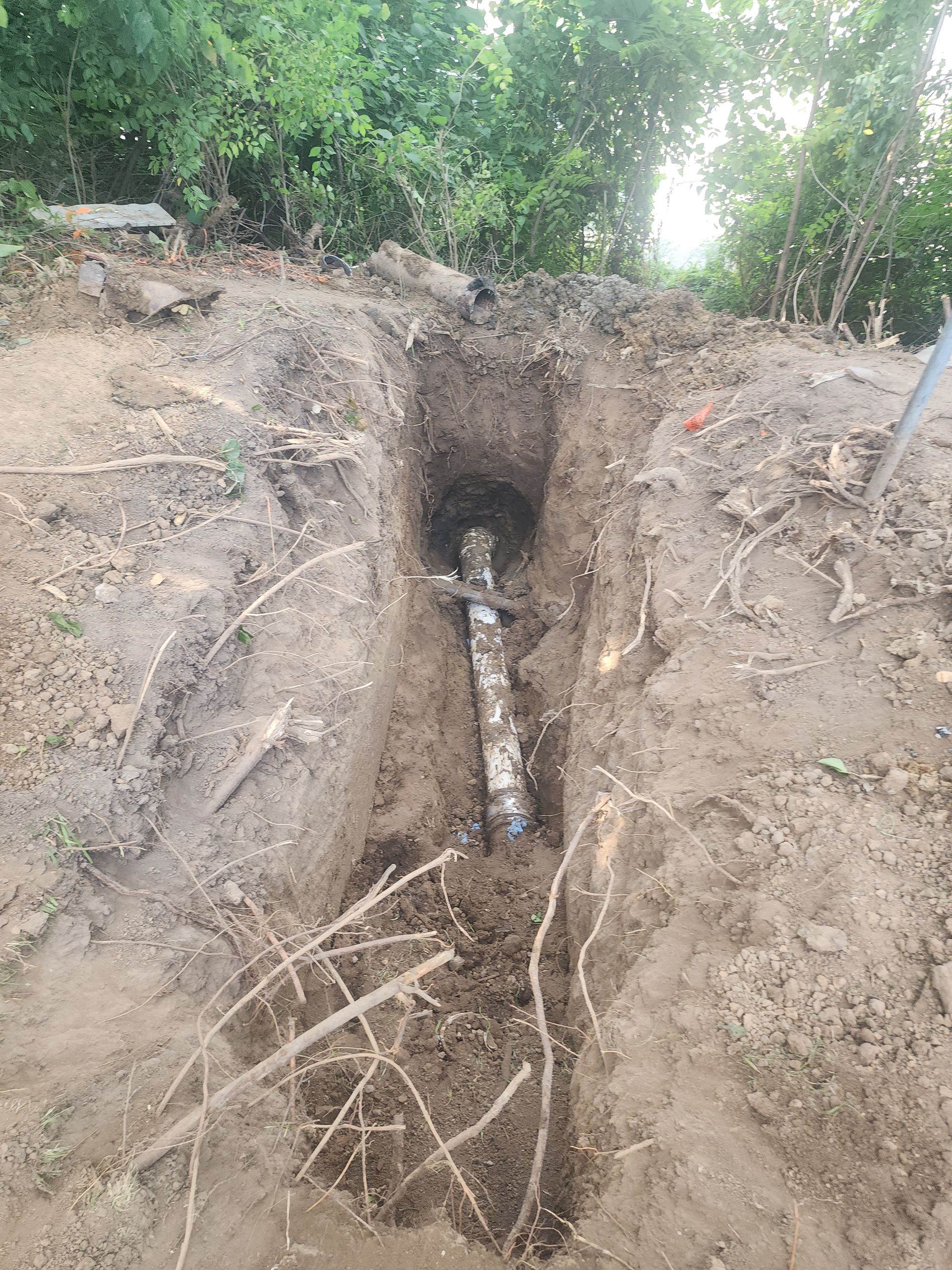 A metallic pipe runs along the bottom of a narrow, freshly dug dirt trench surrounded by trees.