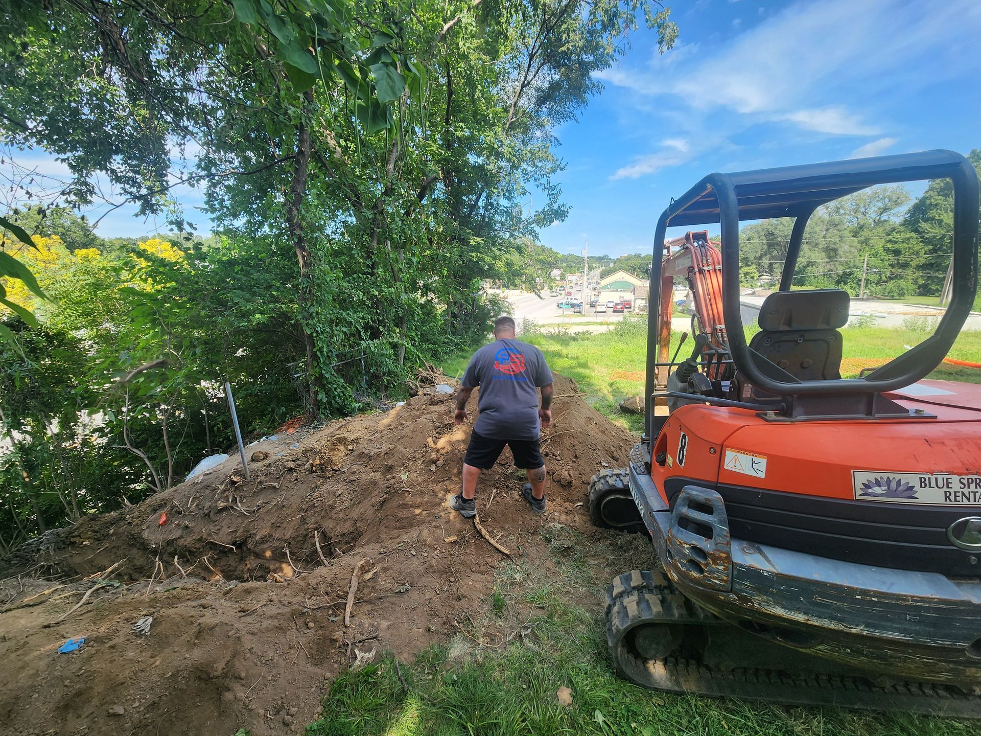 A person in a gray shirt works on a dirt pile next to an orange mini excavator in a grassy, wooded outdoor area.