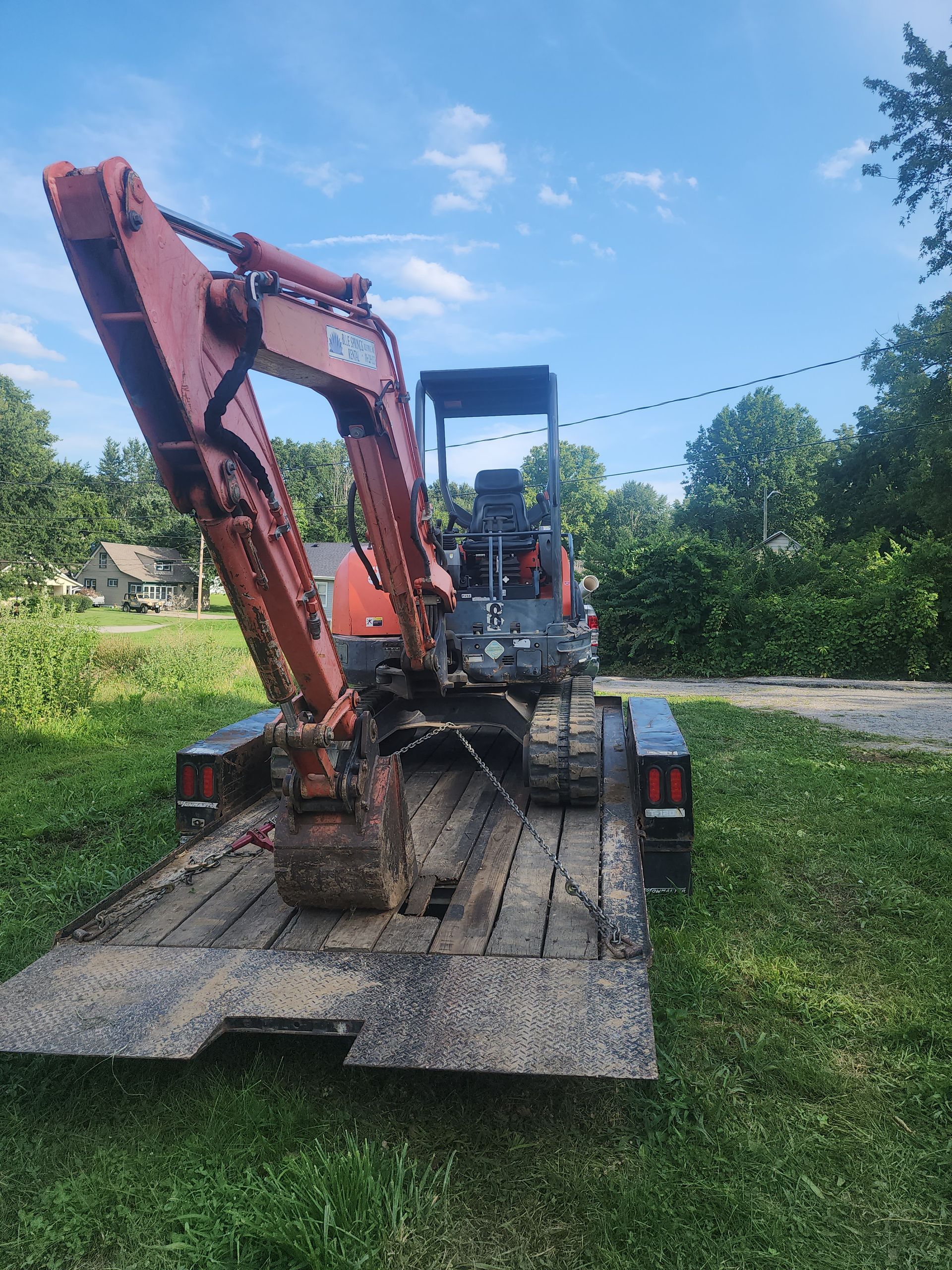 An orange mini-excavator secured on a flatbed trailer outdoors in a grassy, suburban setting under a clear blue sky.