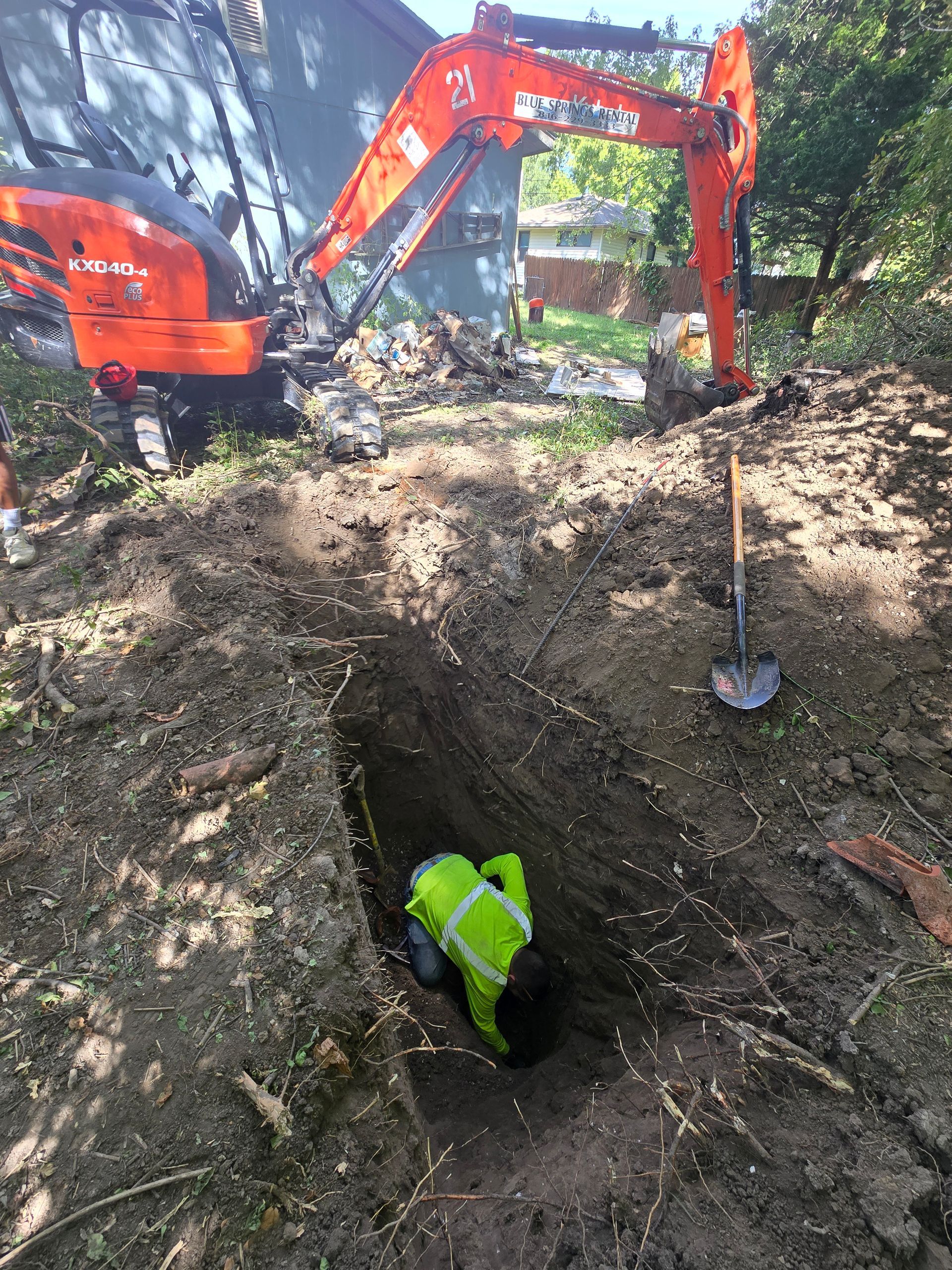 A worker in a high-visibility yellow vest kneels in a deep trench next to an orange Kubota excavator in a wooded area.