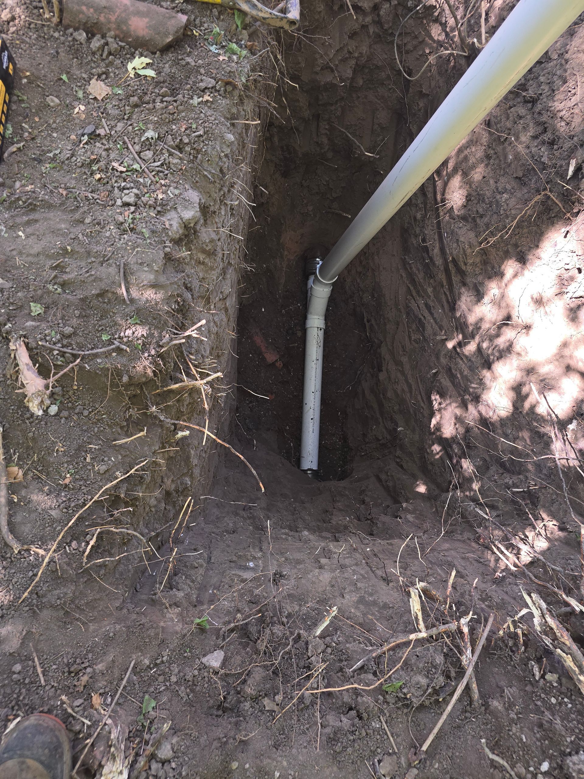 A gray PVC conduit pipe emerges from the bottom of a narrow, freshly dug dirt trench in a wooded area.