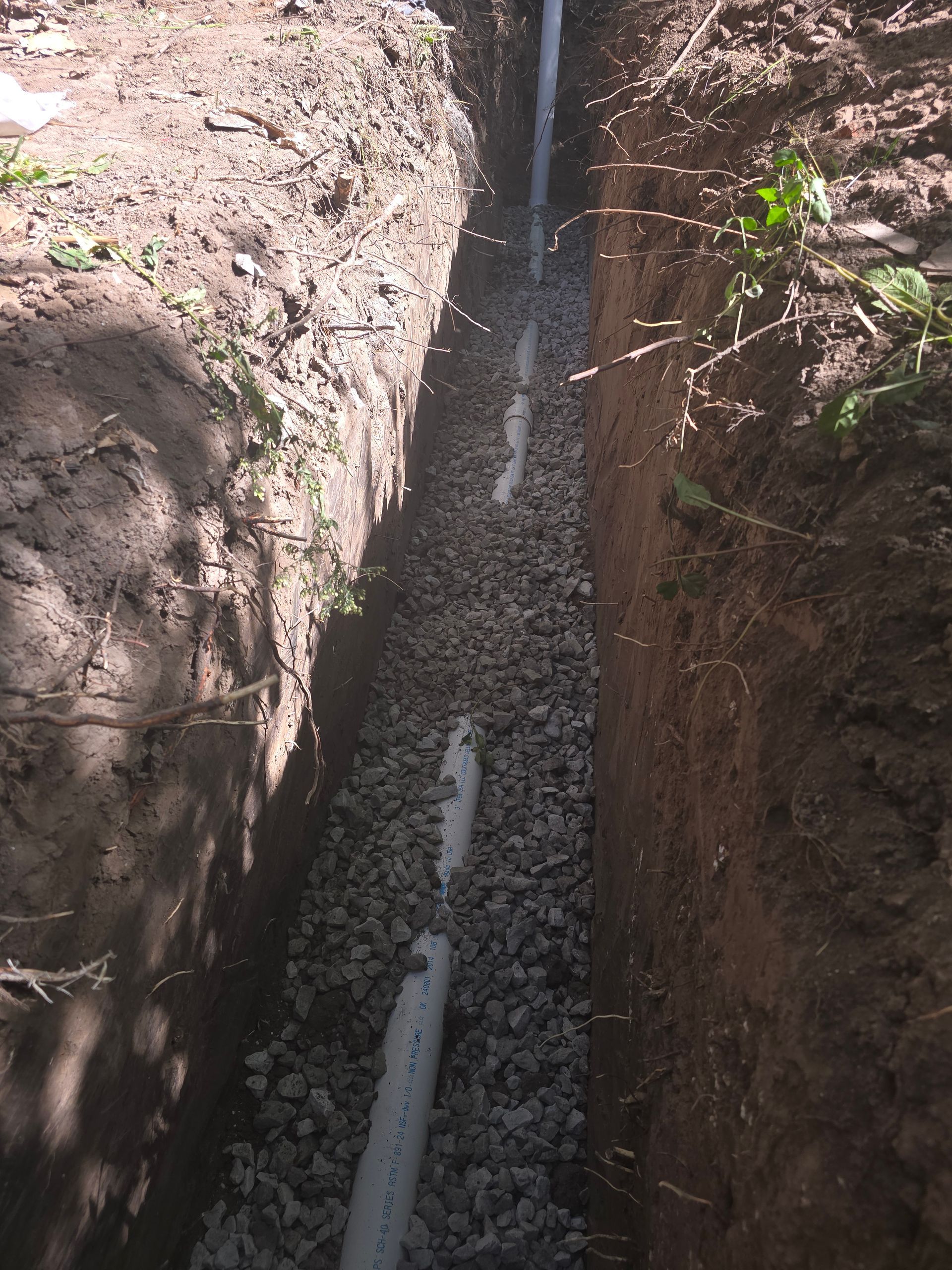 A narrow trench in the ground containing a white PVC pipe laid over a bed of gray gravel.