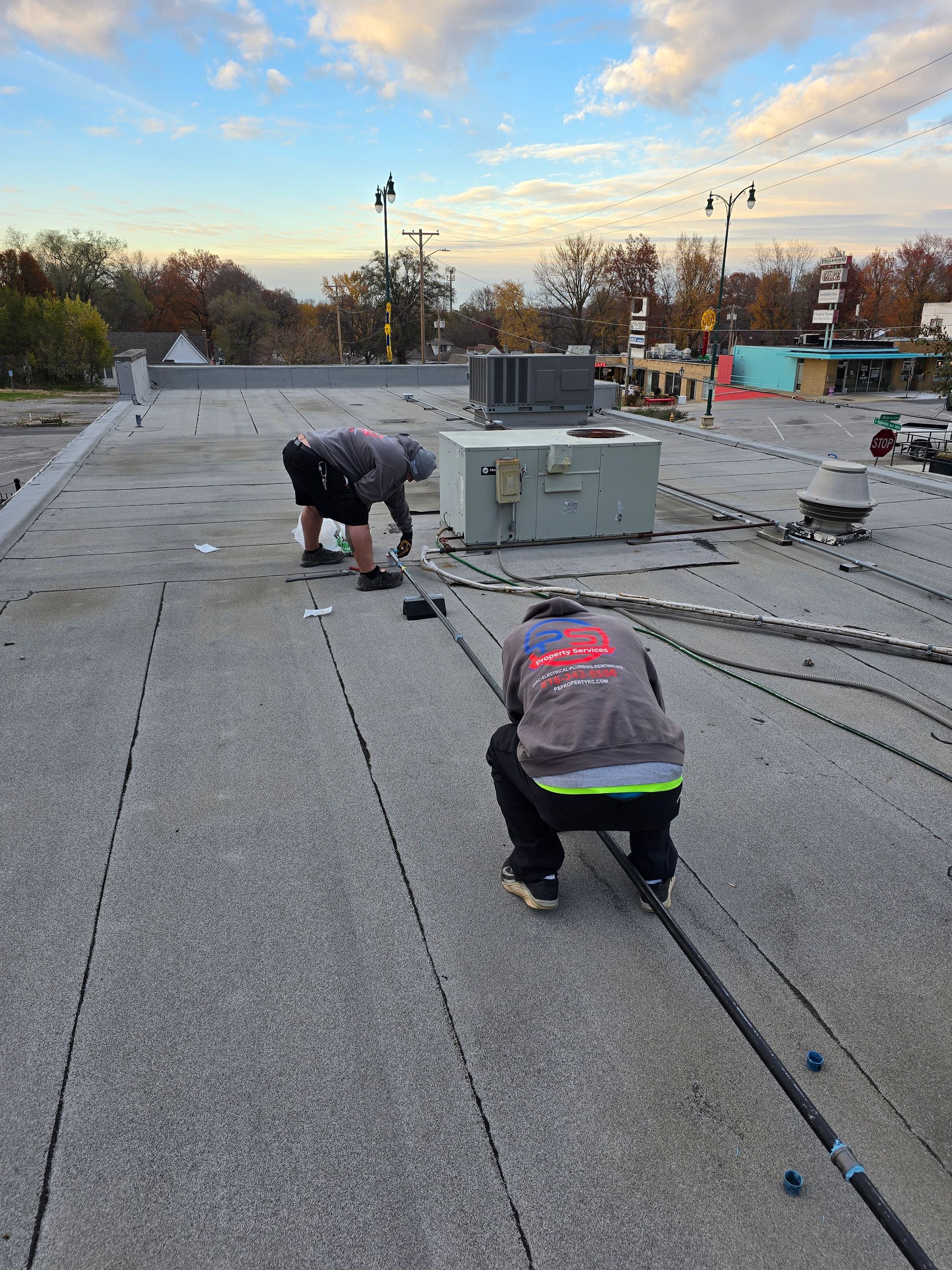 Two workers kneel on a flat gravel roof, performing maintenance work near HVAC units during the late afternoon.