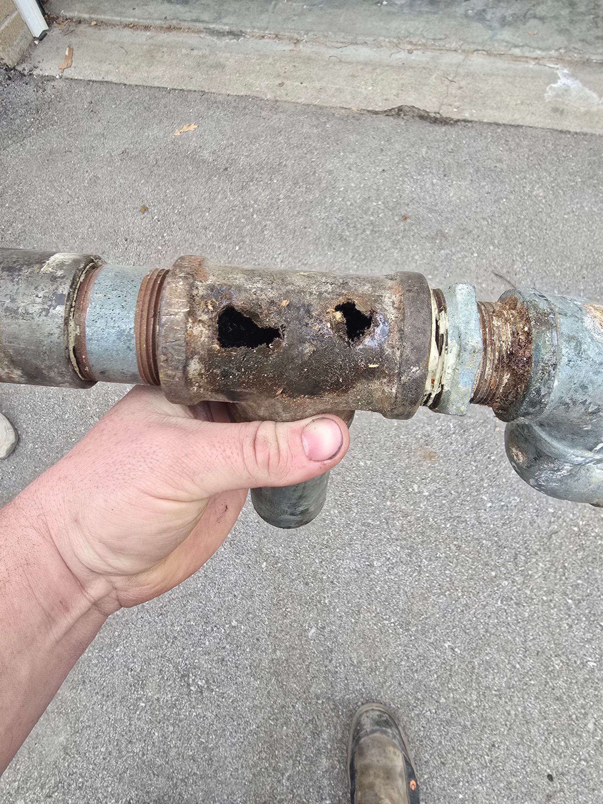 A hand holds a severely corroded and rusted iron pipe fitting with two large holes in it.