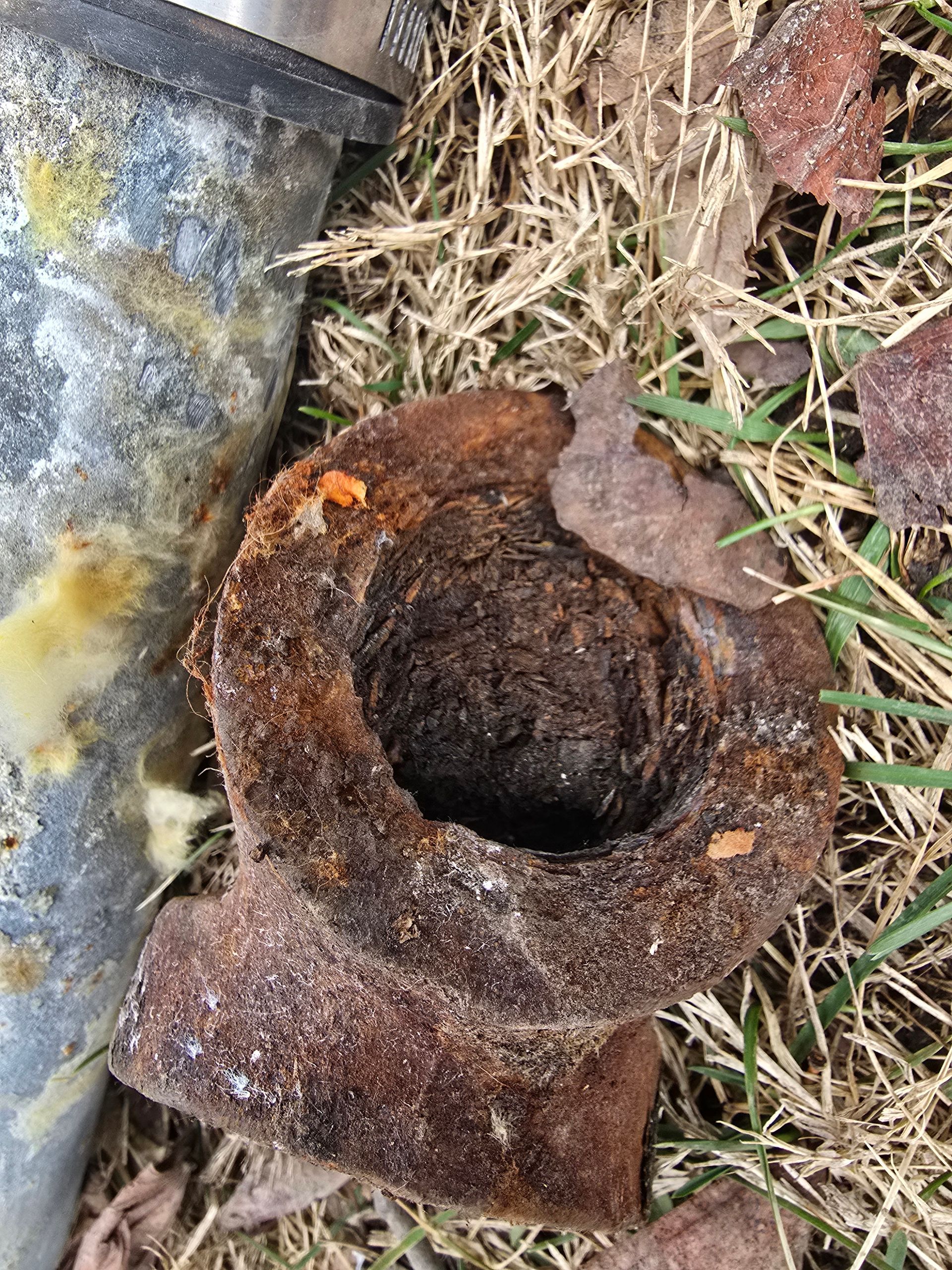 A close-up of a heavily rusted, circular metal pipe fitting lying on dead grass near a concrete surface.