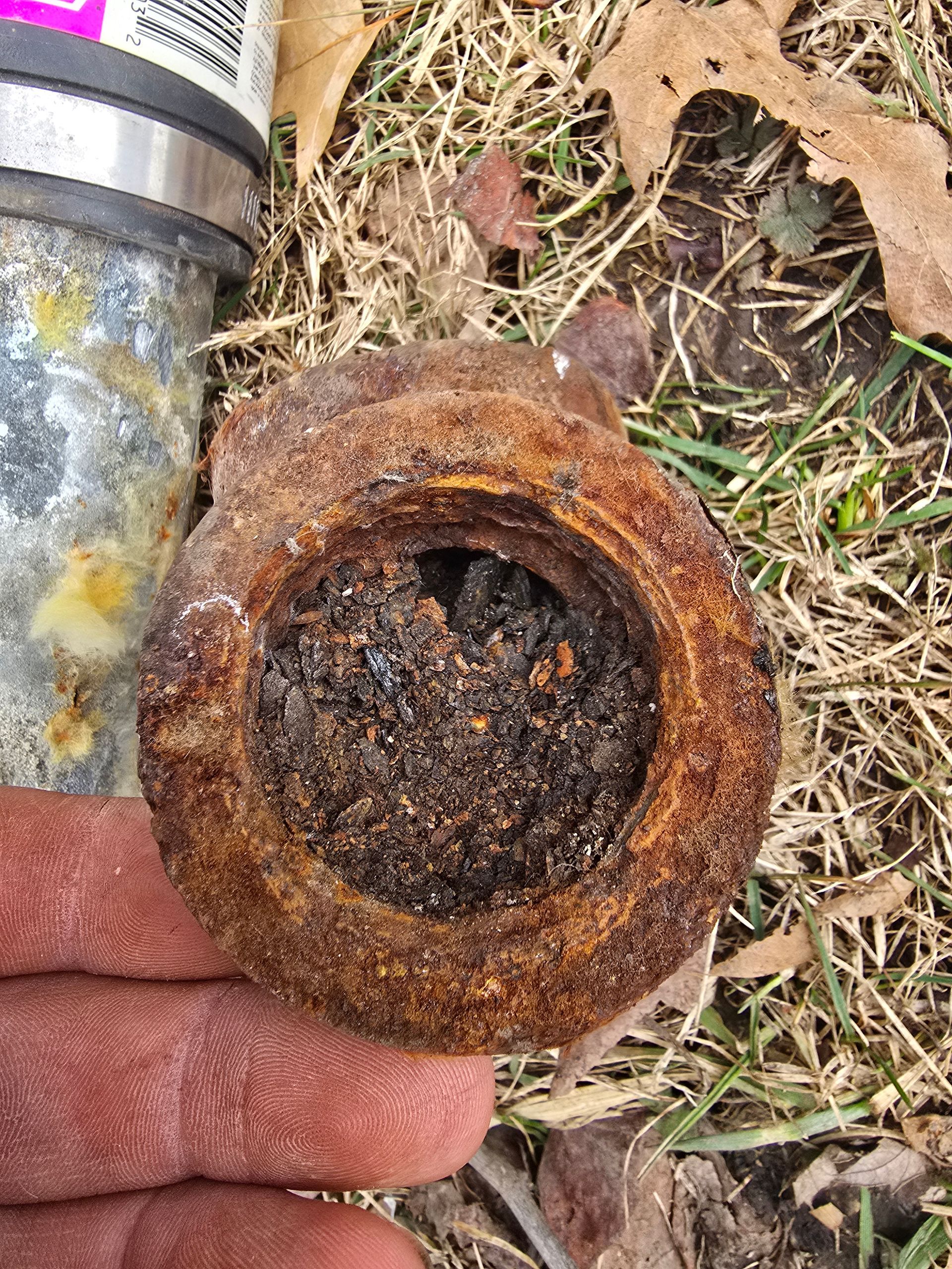 A gloved hand holds a heavily rusted, circular metal pipe fitting, which is filled with dark, loose debris.