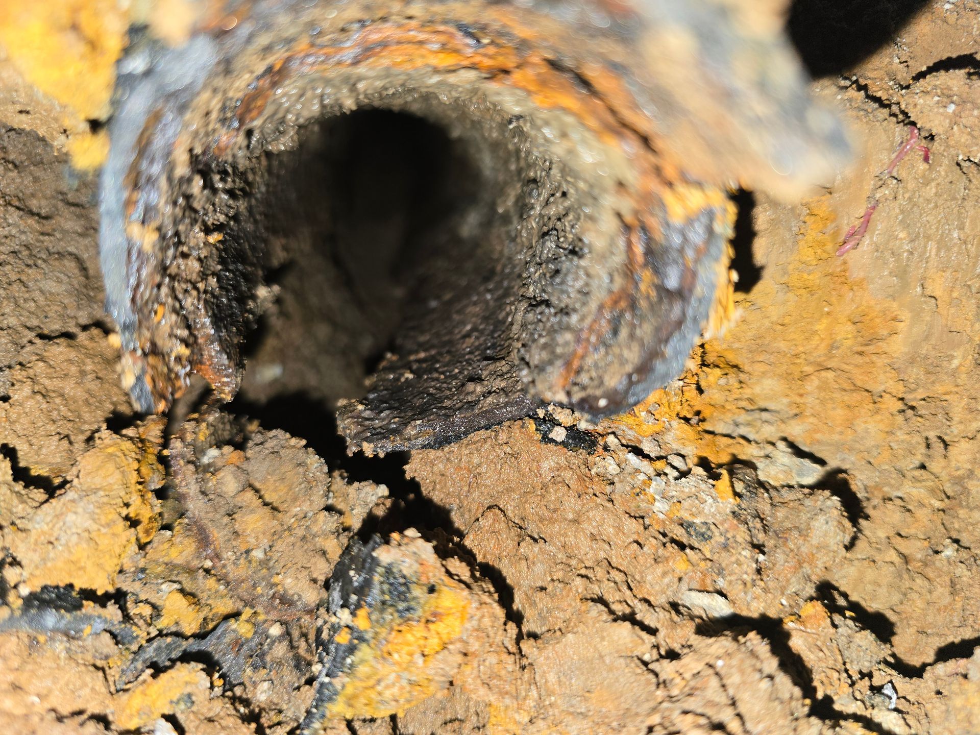 A close-up view looking into the end of a corroded, debris-filled metal pipe surrounded by dry, cracked soil.