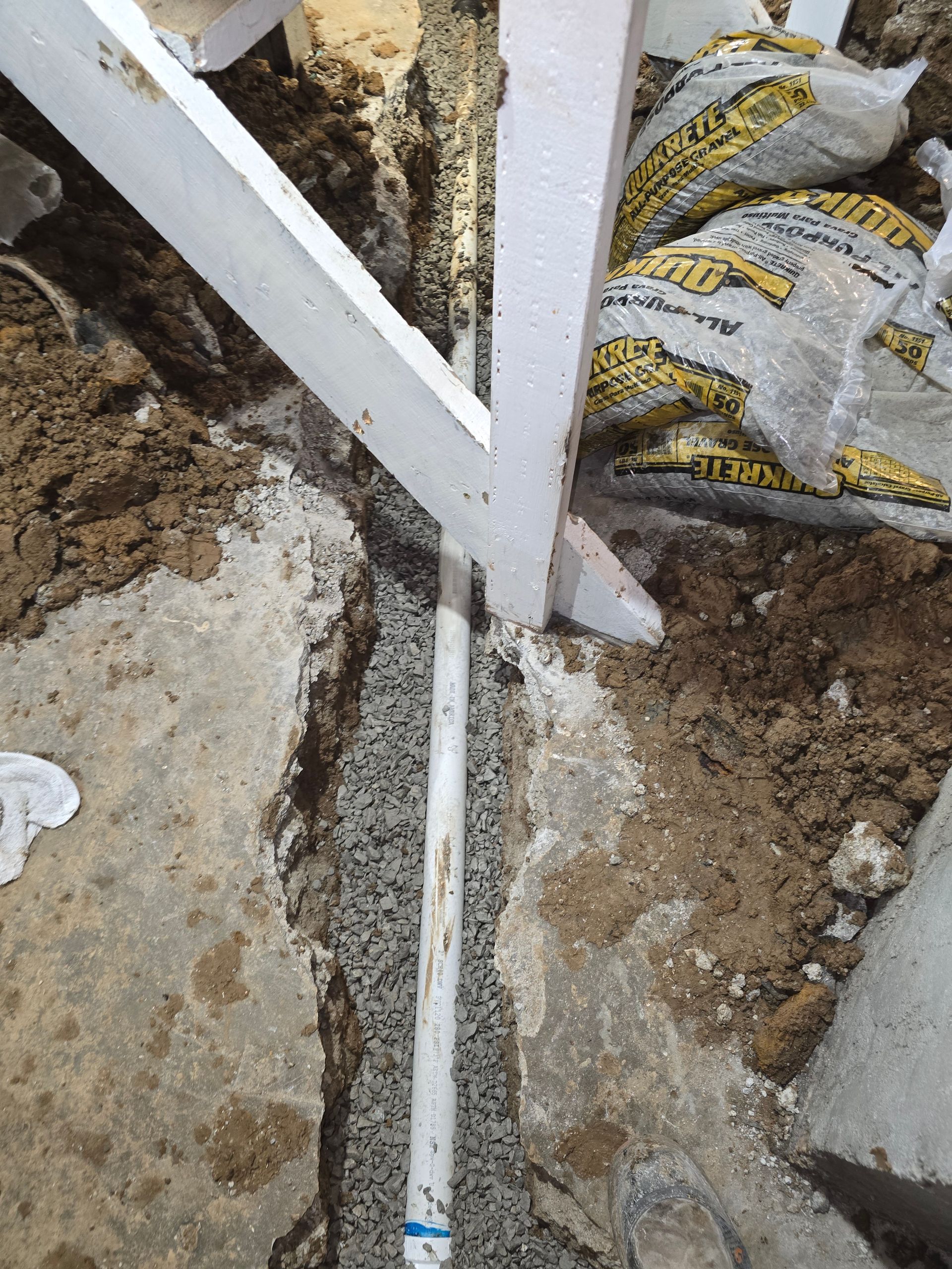 A white pipe lies in a gravel-filled trench cut through concrete, positioned beneath wooden stairs.