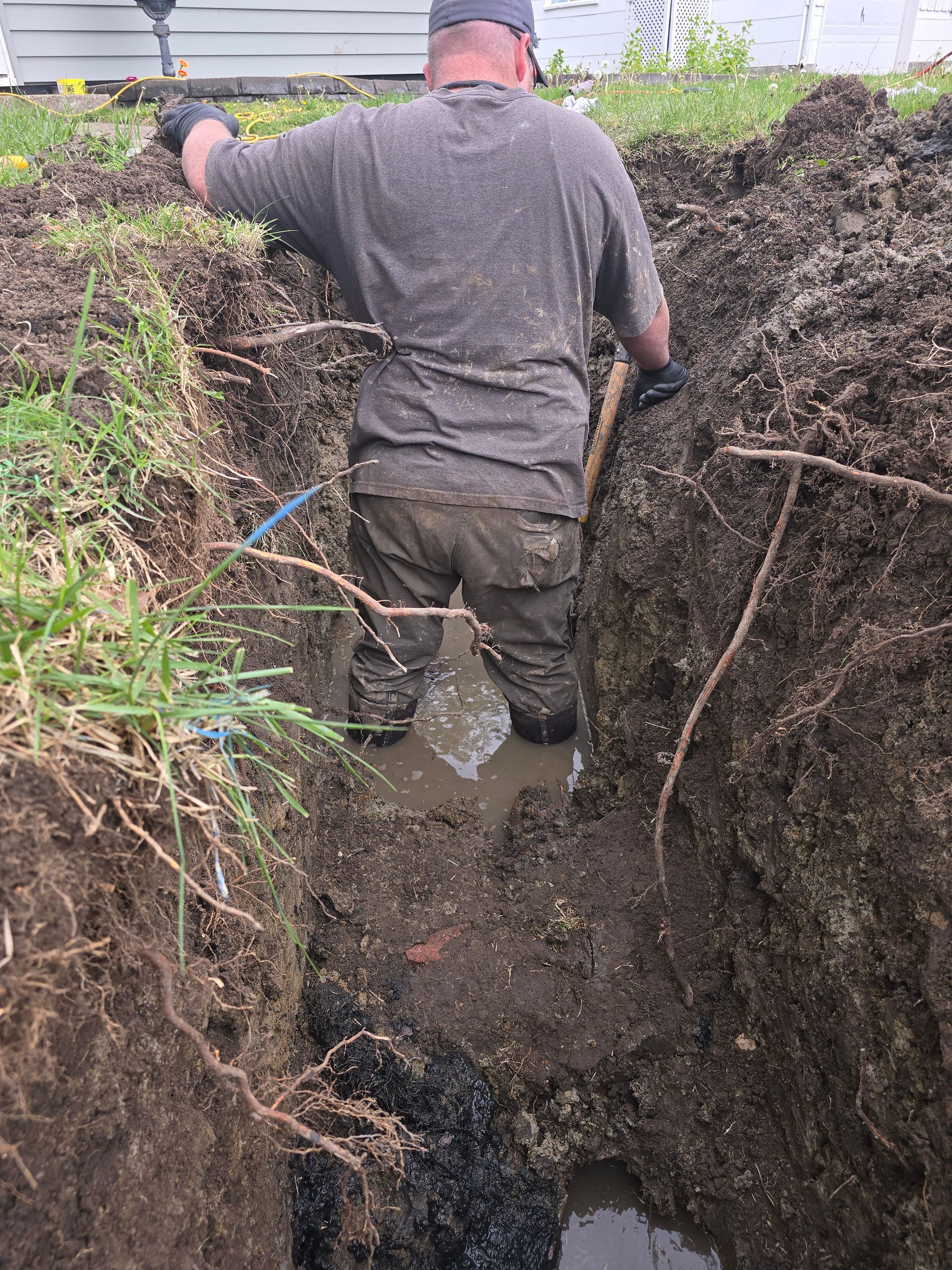 A person wearing a grey shirt and dark pants stands knee-deep in a muddy trench, working on a project in an outdoor area.