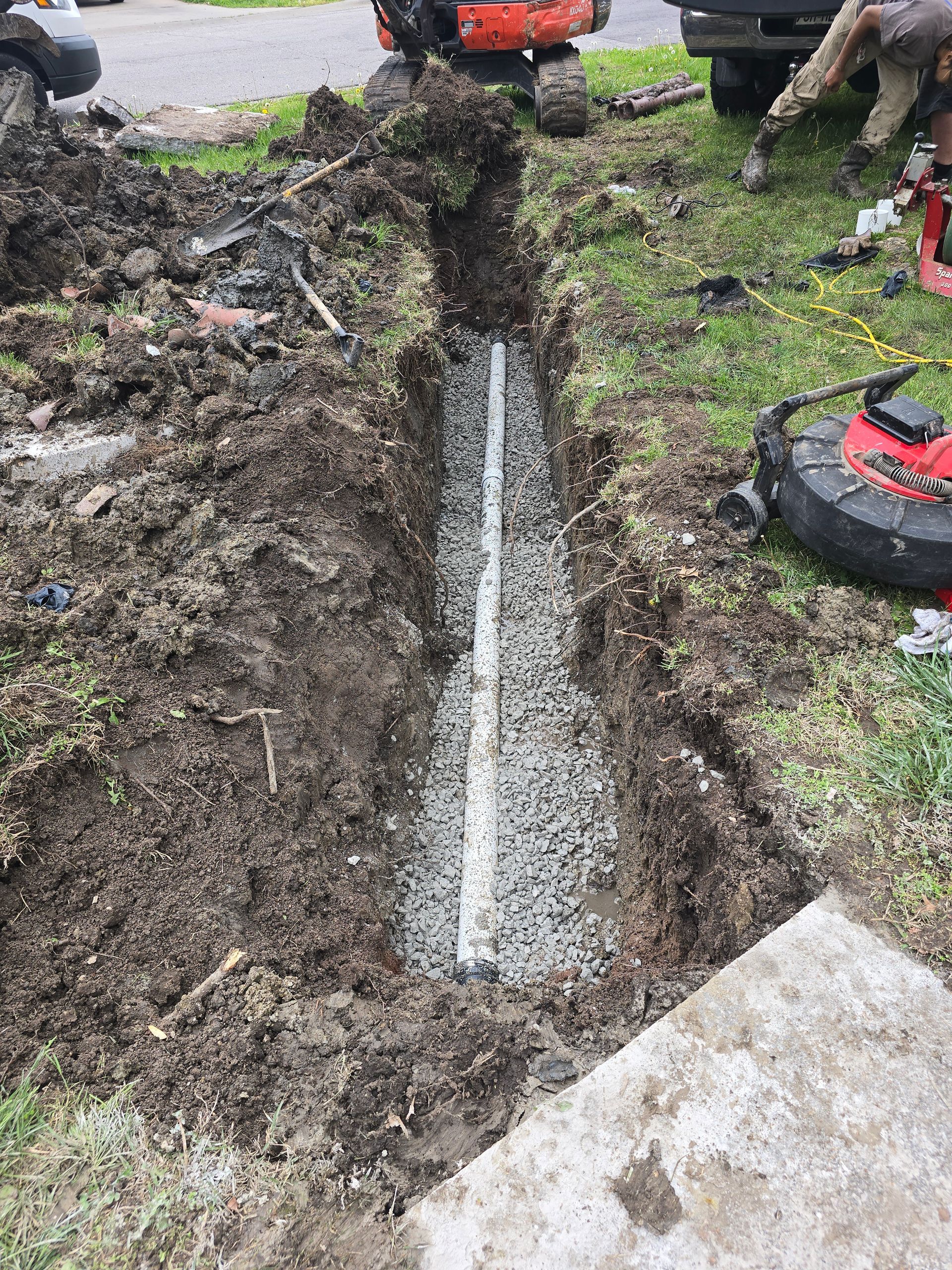 A pipe sits in a trench filled with gravel, with a lawnmower, construction machinery, and soil visible nearby.