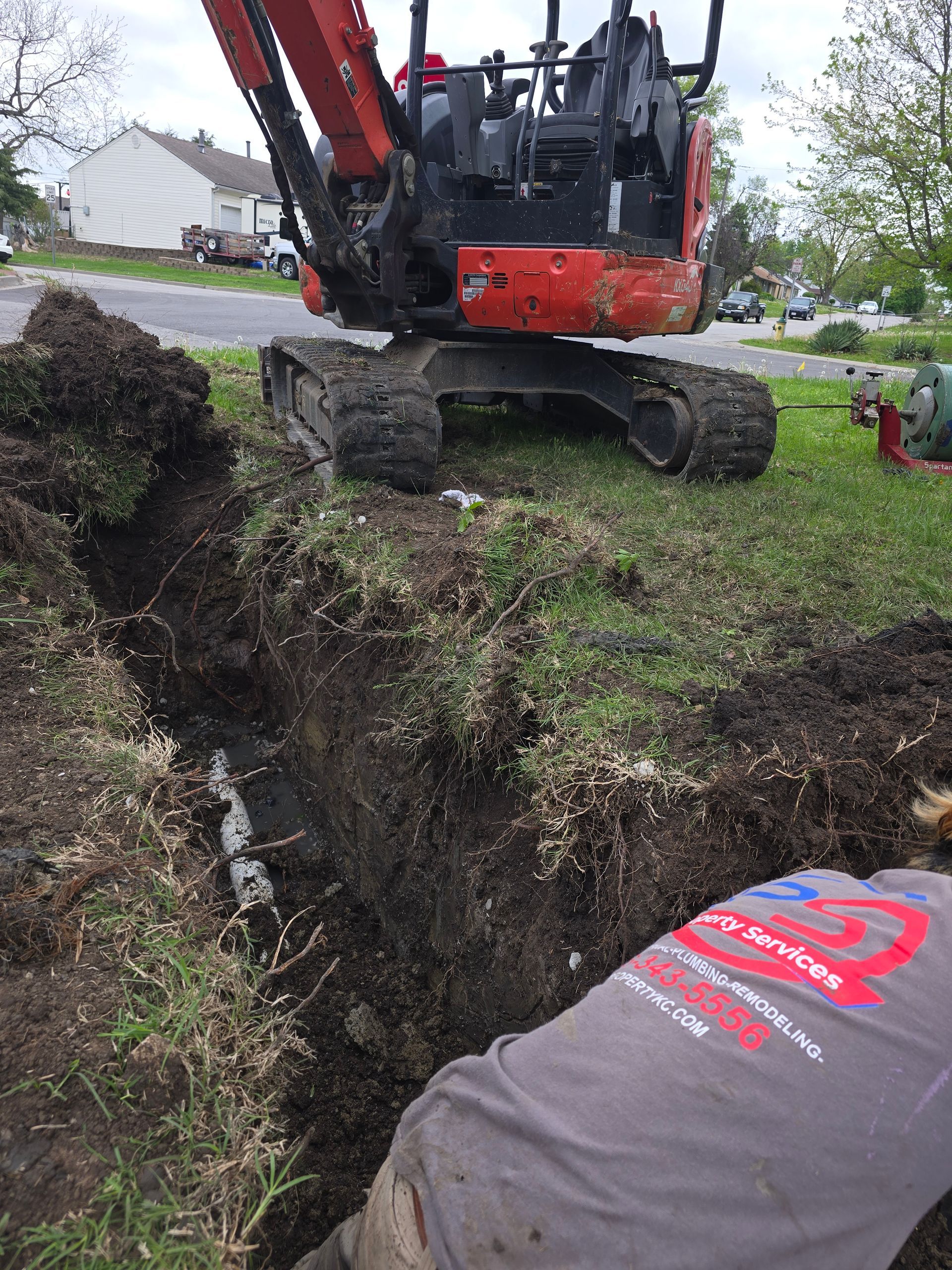 A person in a work shirt kneels by a trench in a grassy yard while a red excavator sits behind them.