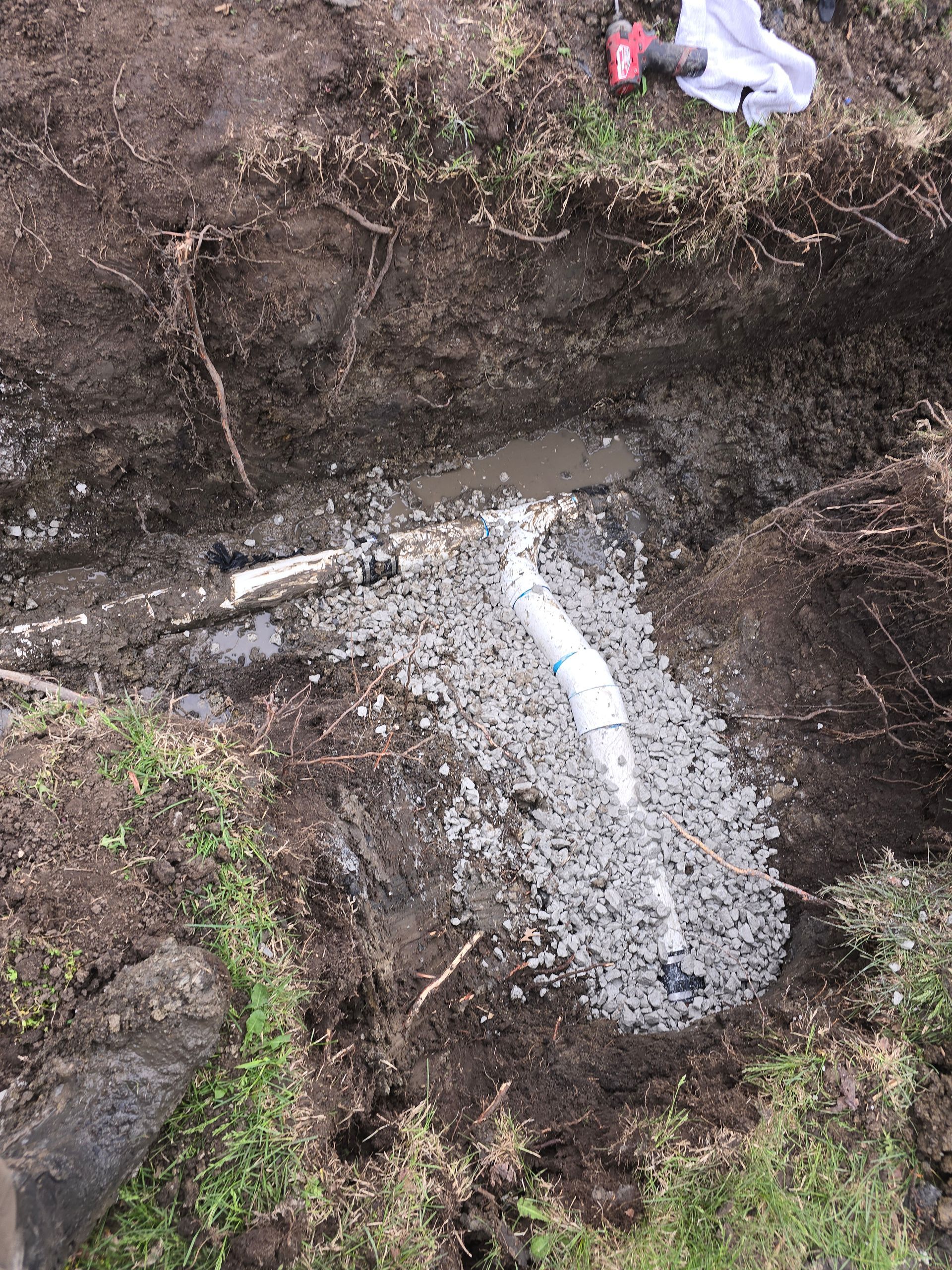 A white PVC pipe joined by a T-connector sits in a muddy trench filled with grey gravel.