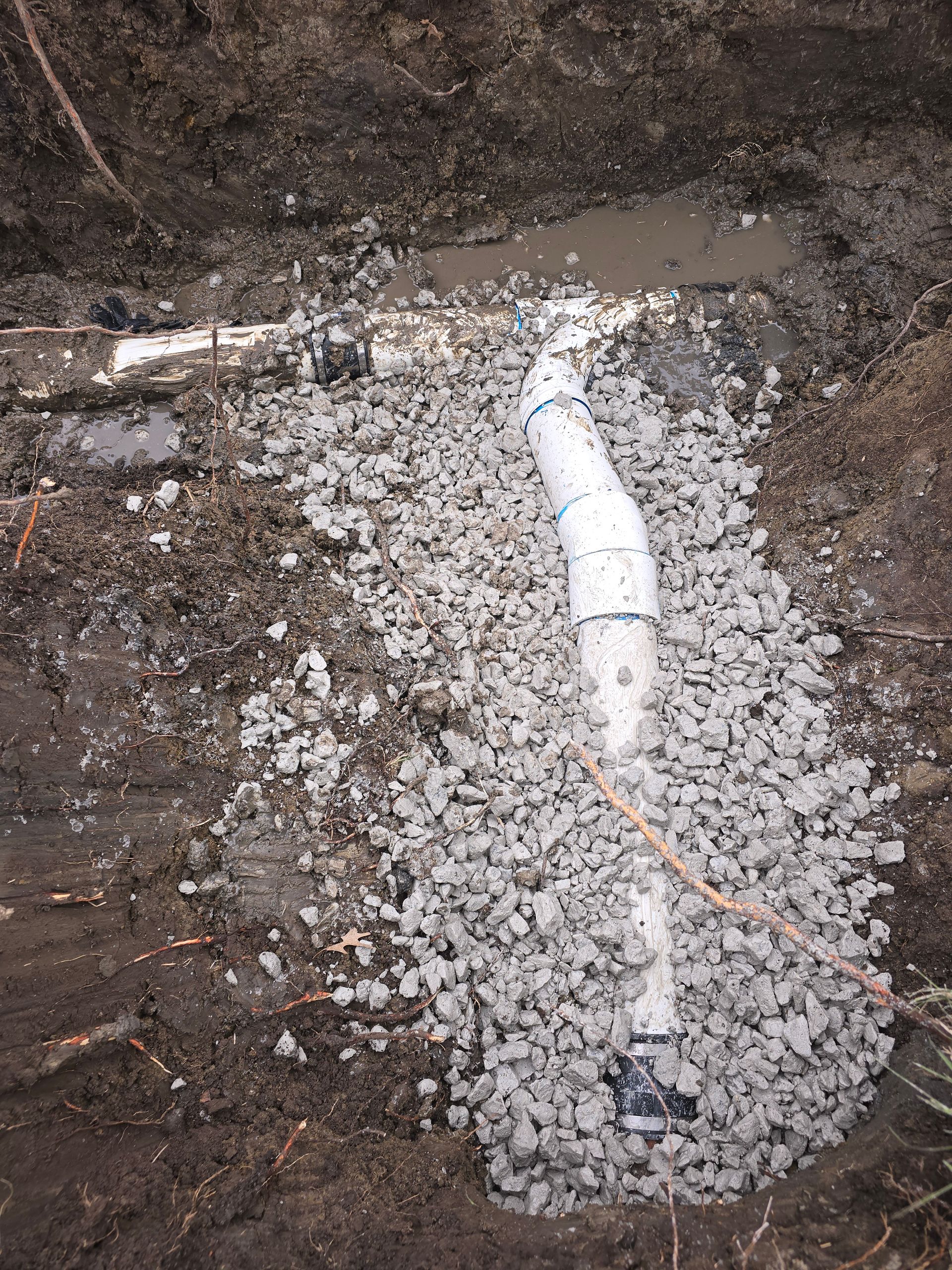 A white PVC pipe assembly with multiple joints rests in a dirt trench, partially covered by grey gravel.