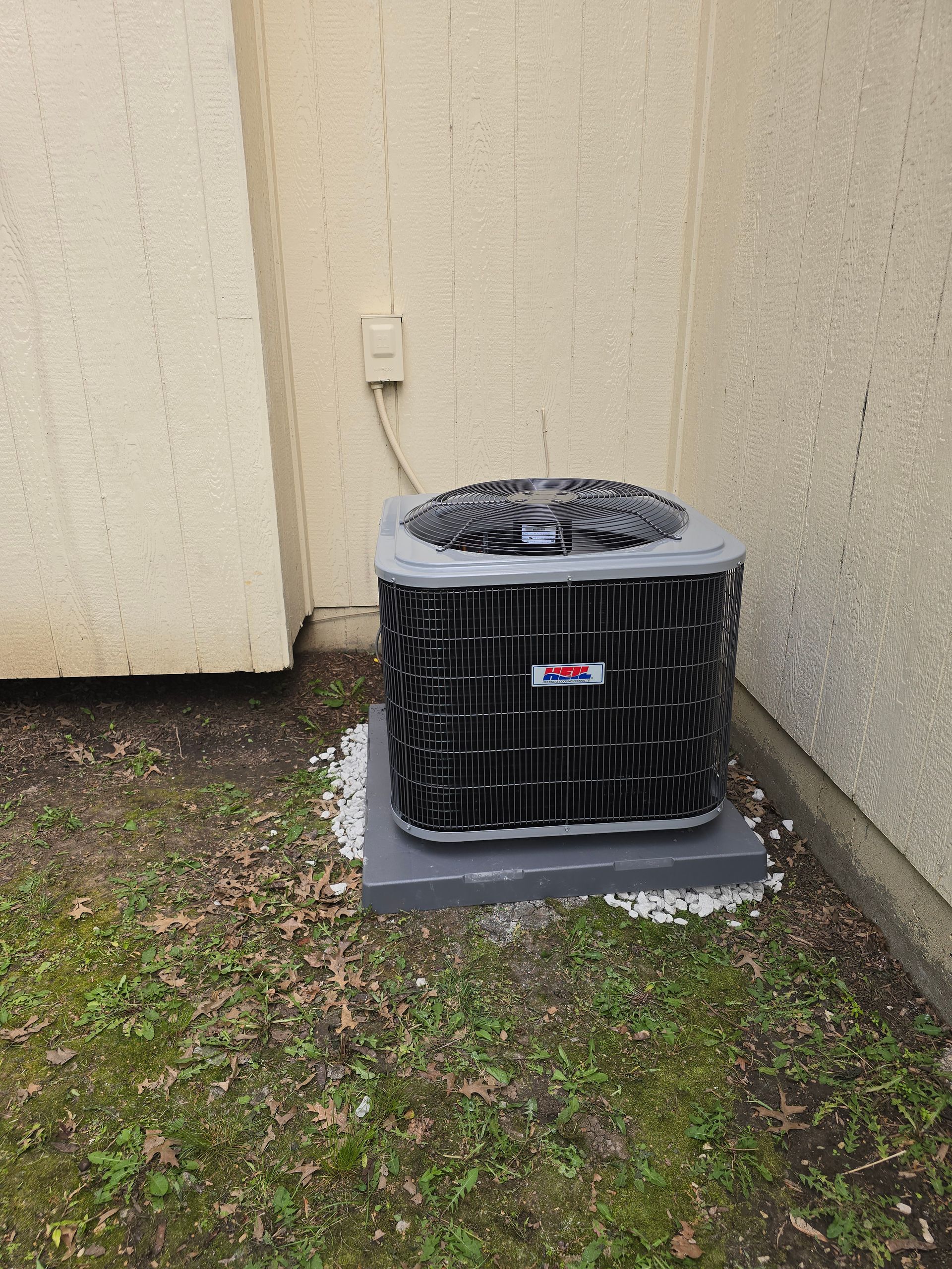 A gray HVAC air conditioning condenser unit sitting on a concrete pad in the corner of a tan exterior house wall.