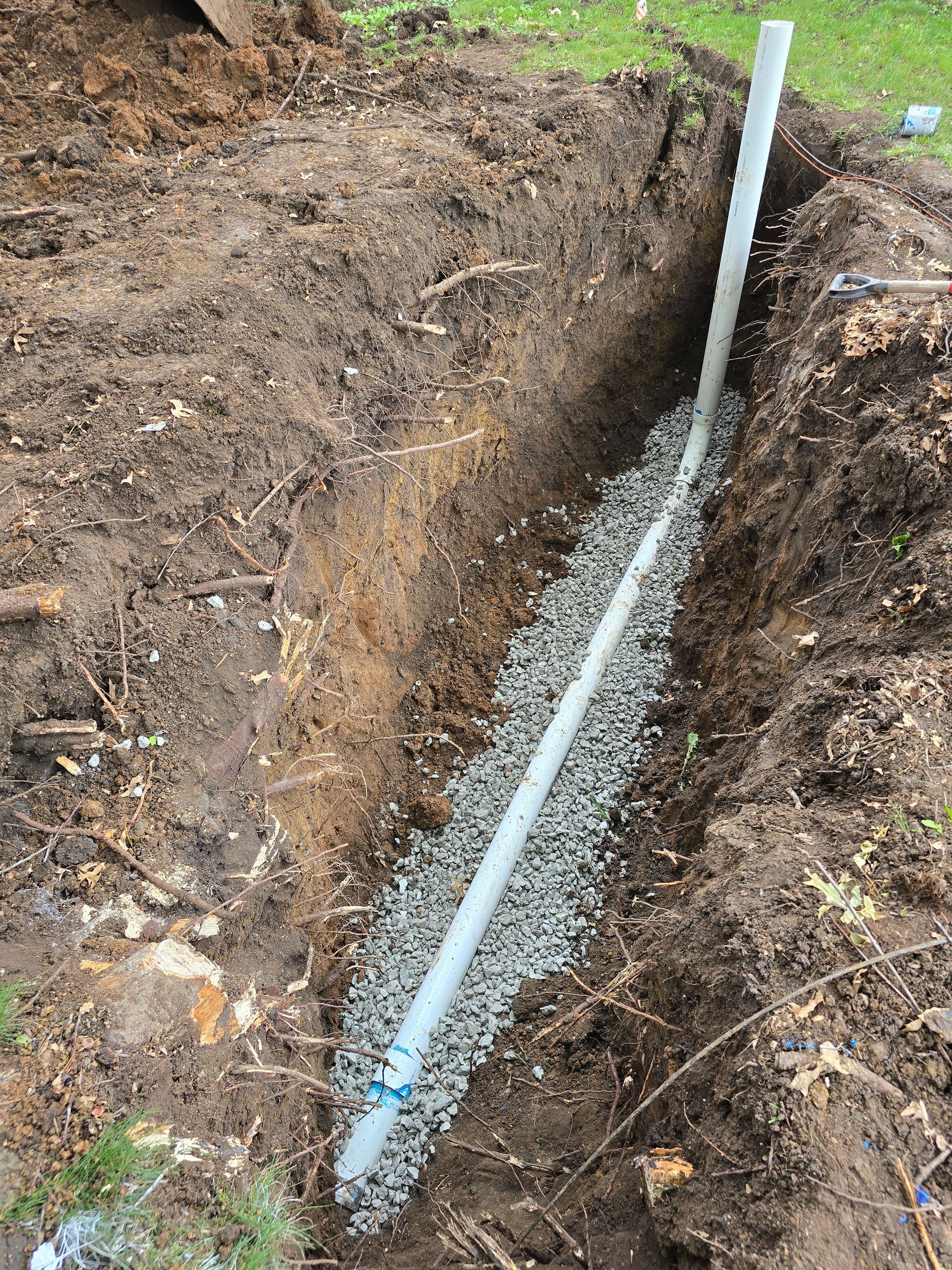 A white perforated PVC drainage pipe sits inside a narrow, gravel-lined trench in the dirt.