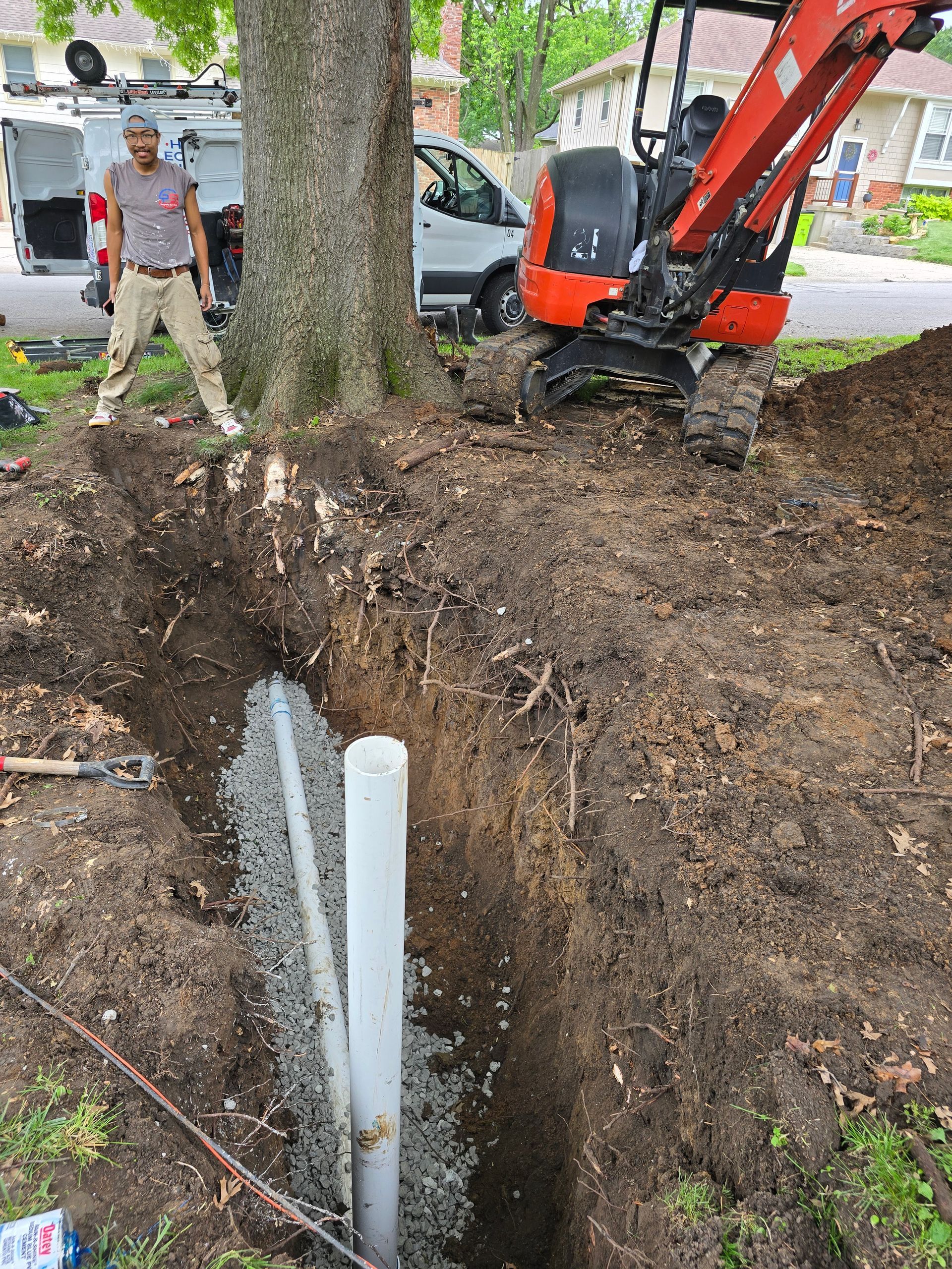 A worker stands near a dug trench containing a vertical white pipe and a gravel-filled drainage line near a large tree.