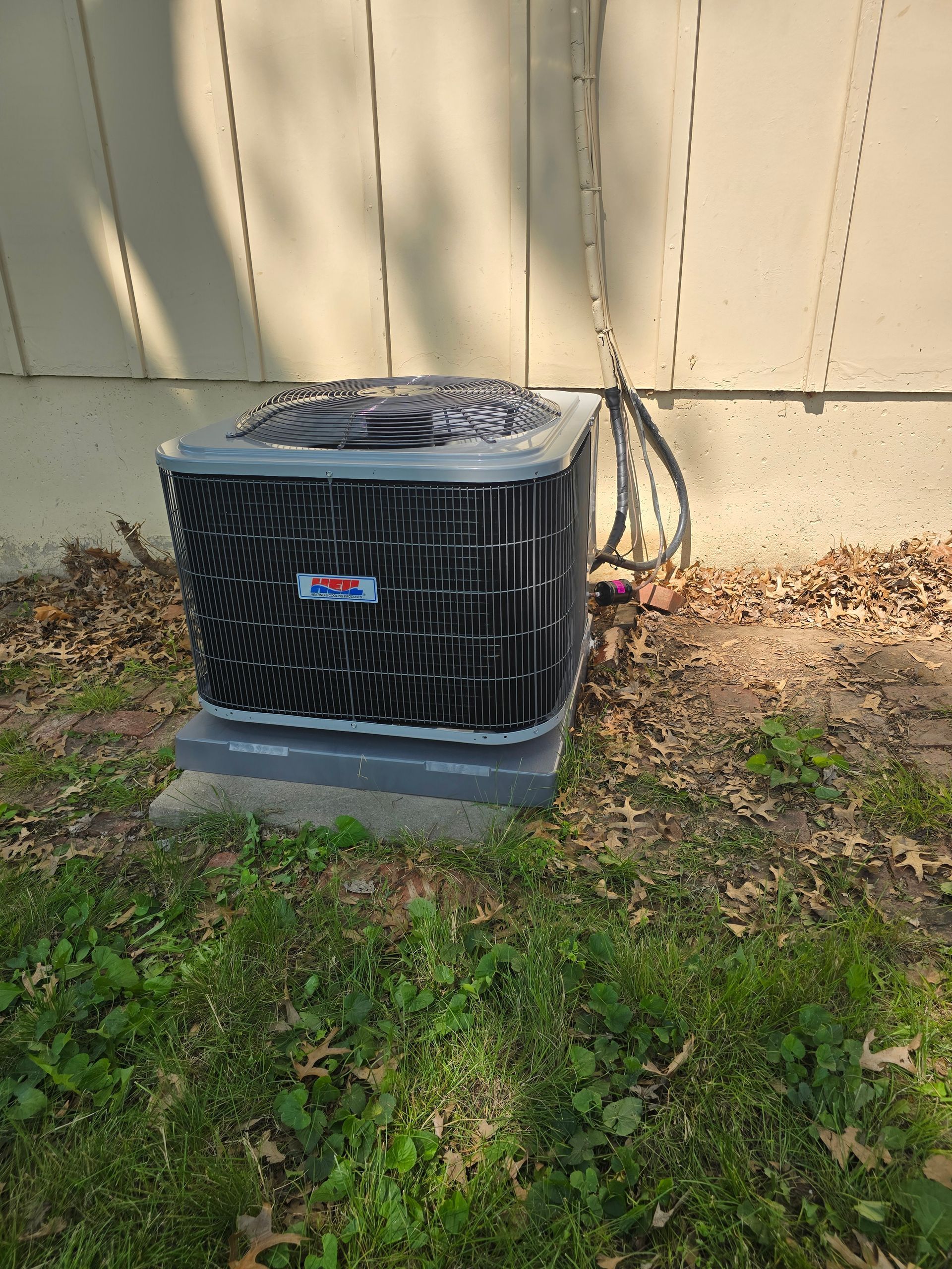 A residential air conditioning unit sitting on a concrete pad next to an exterior house wall on a grassy lawn.