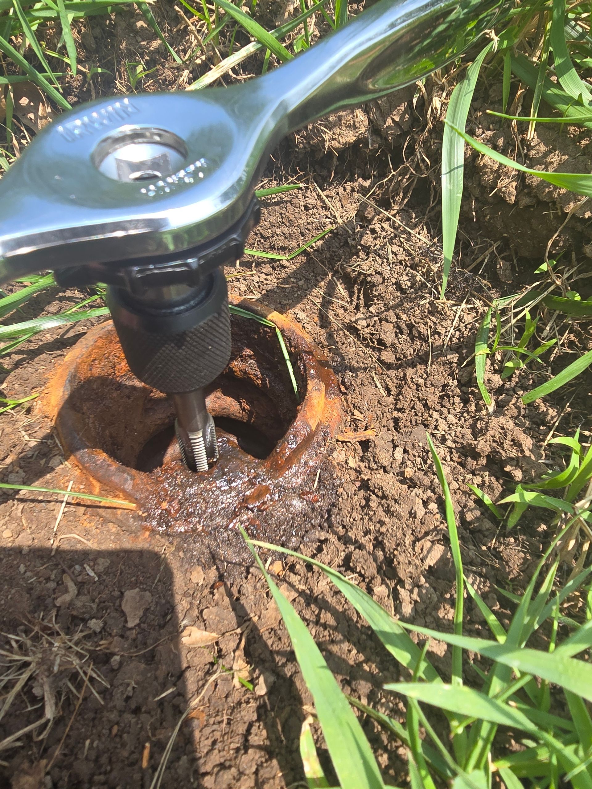 A metal ratcheting tool inserted into a rusted, circular valve access pipe embedded in the ground.