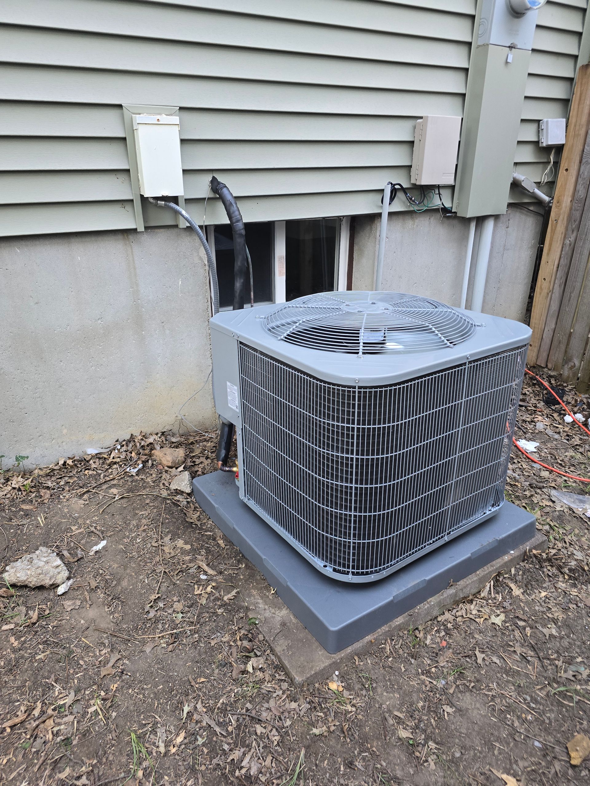 A gray central air conditioning unit sits on a concrete pad against the light-colored siding of a house foundation.