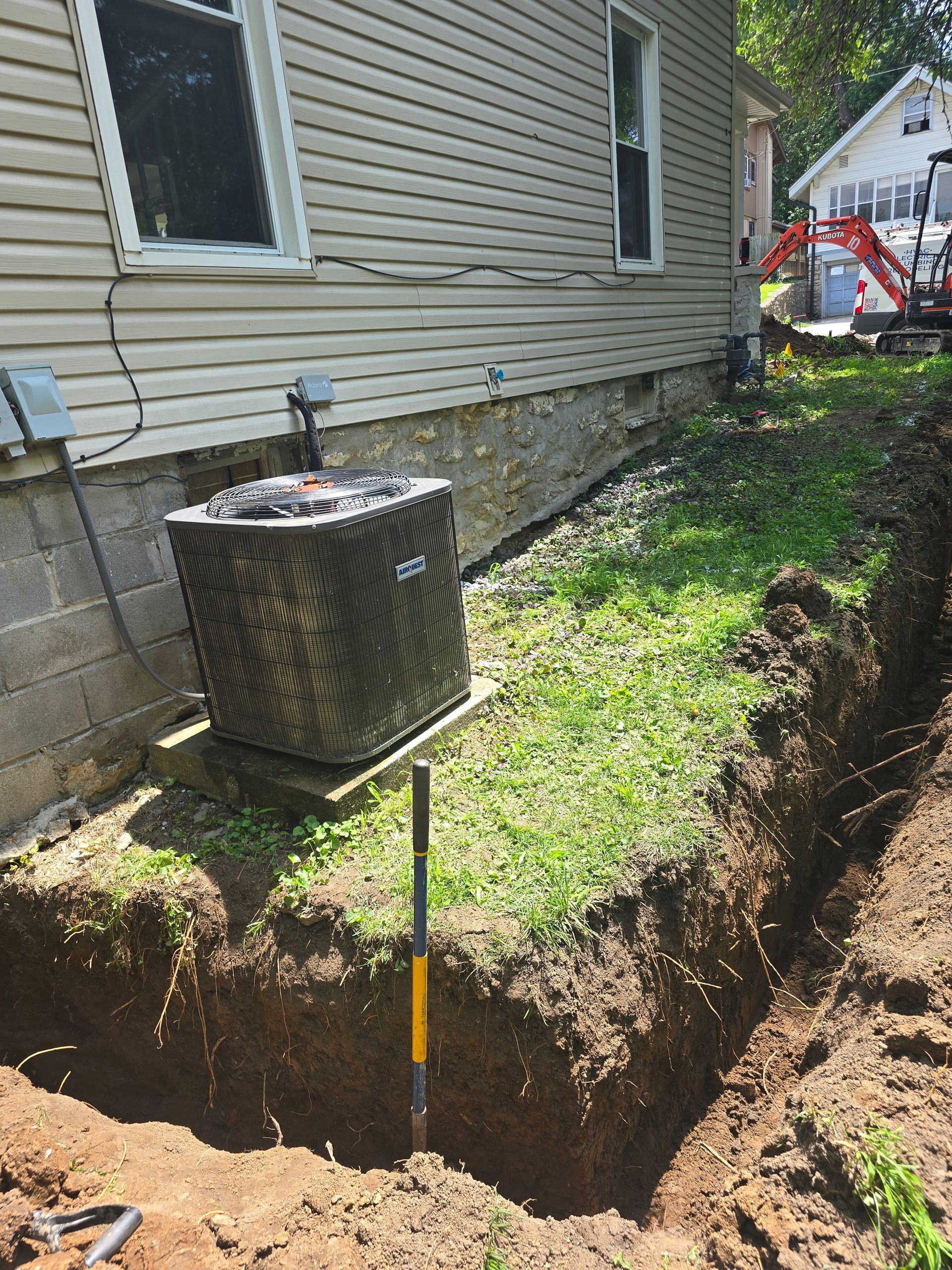 A trench is dug beside a house foundation, near an AC unit and some construction equipment in the background.