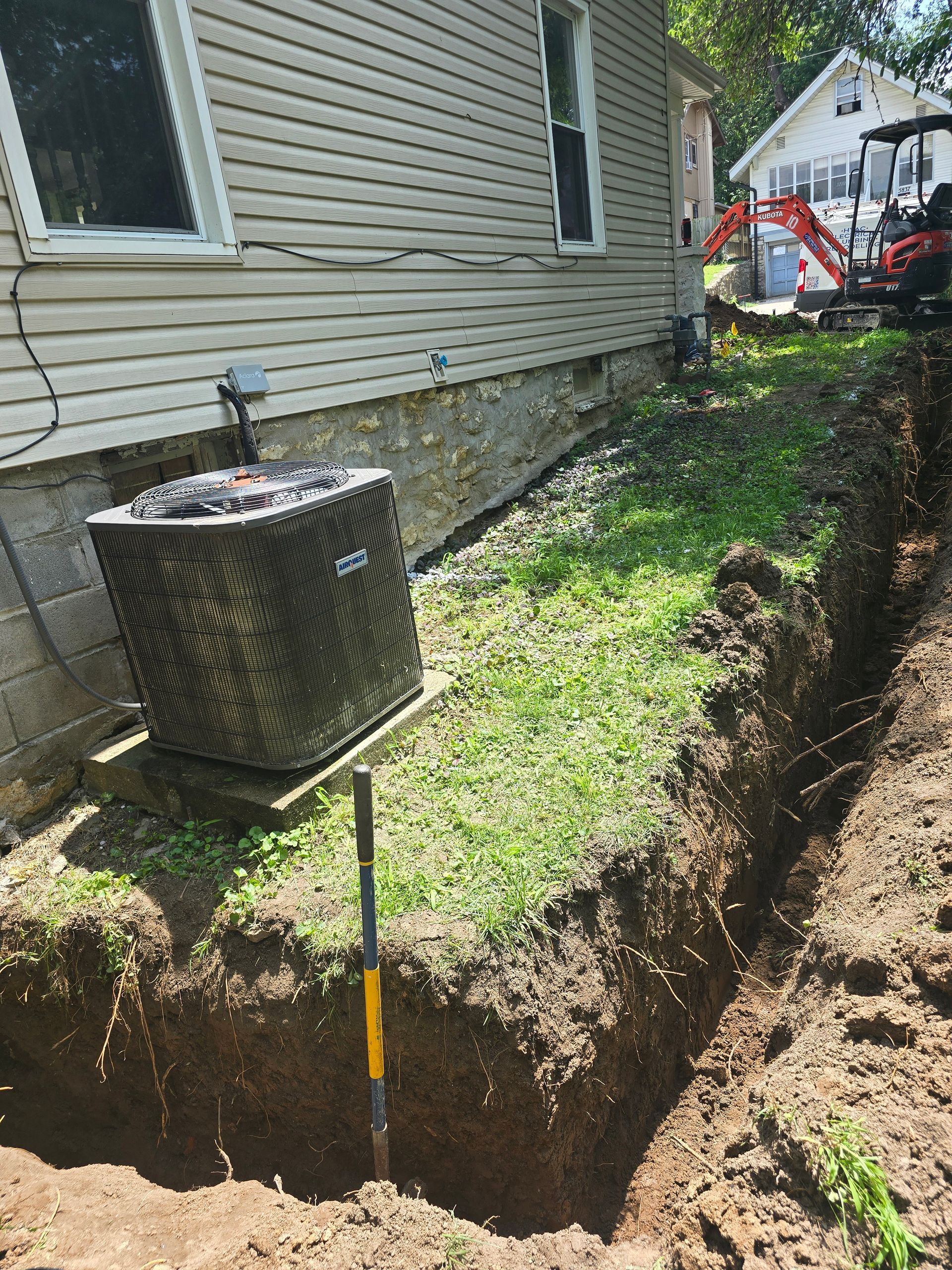 A side view of a house foundation with an air conditioner unit next to a deep excavation trench in the yard.