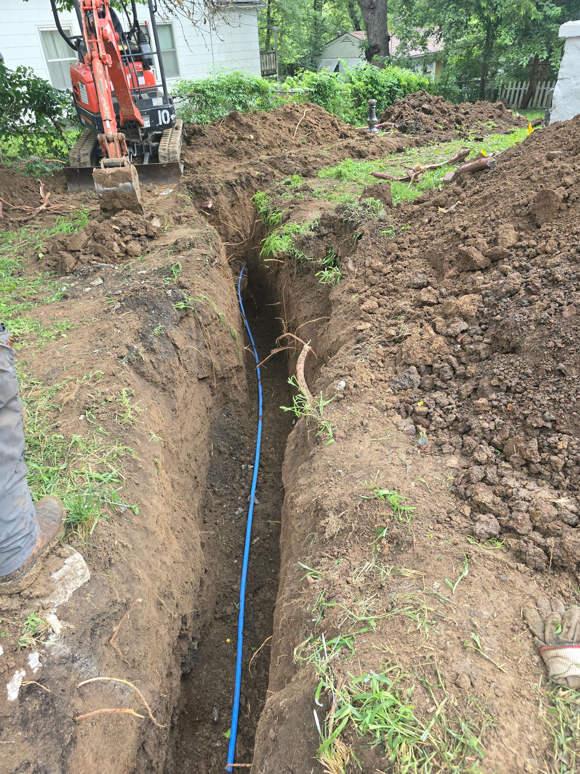 An orange mini-excavator sits by a dirt trench containing a blue pipe in a residential yard.