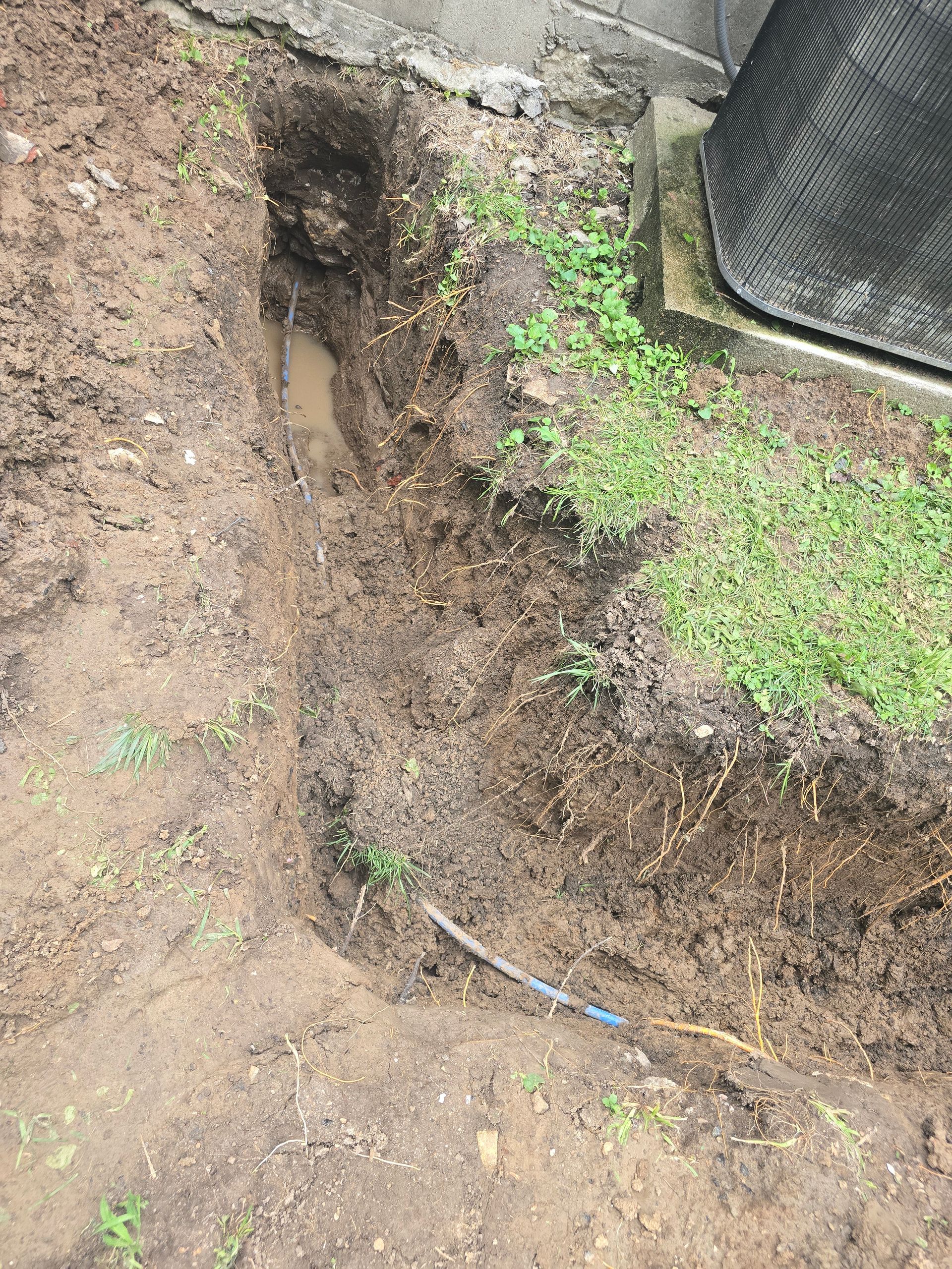 A muddy trench dug into the ground next to a concrete wall and a black HVAC unit, with a blue pipe visible at the bottom.