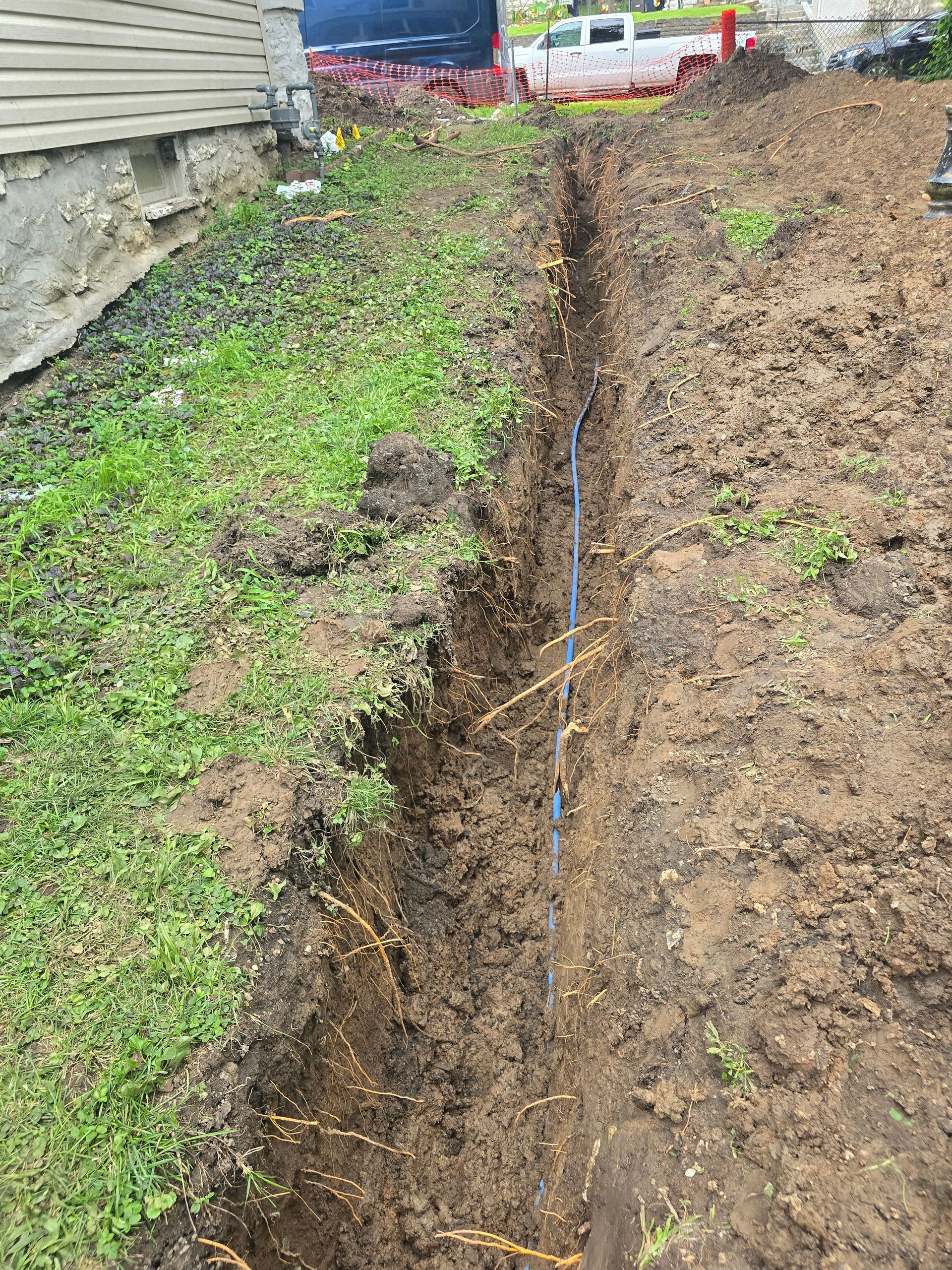 A long, narrow trench dug in the ground next to a house foundation, showing a blue pipe running through the center.
