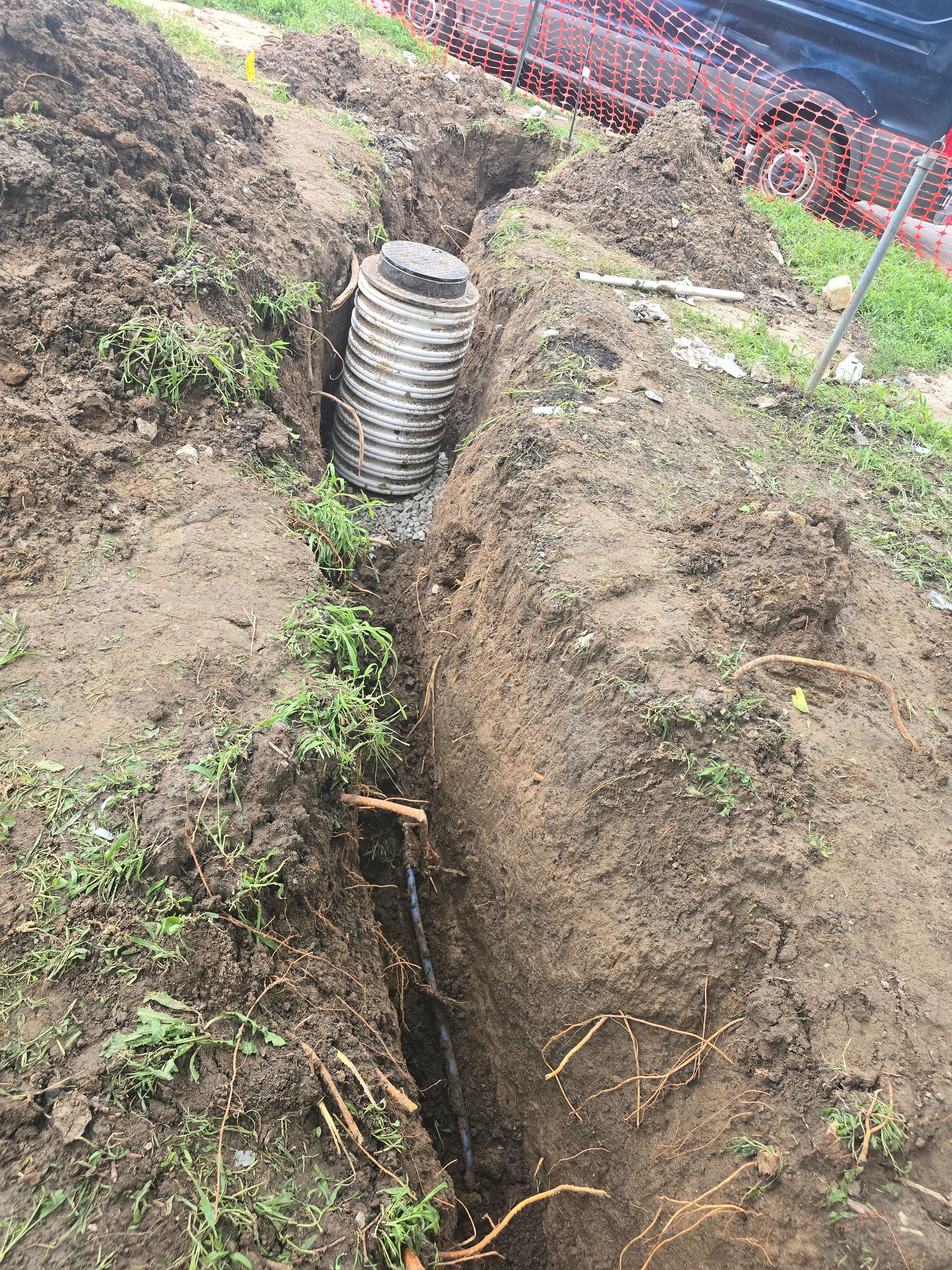 A view from above showing a circular black drainage basin installed in a narrow, freshly dug dirt trench.