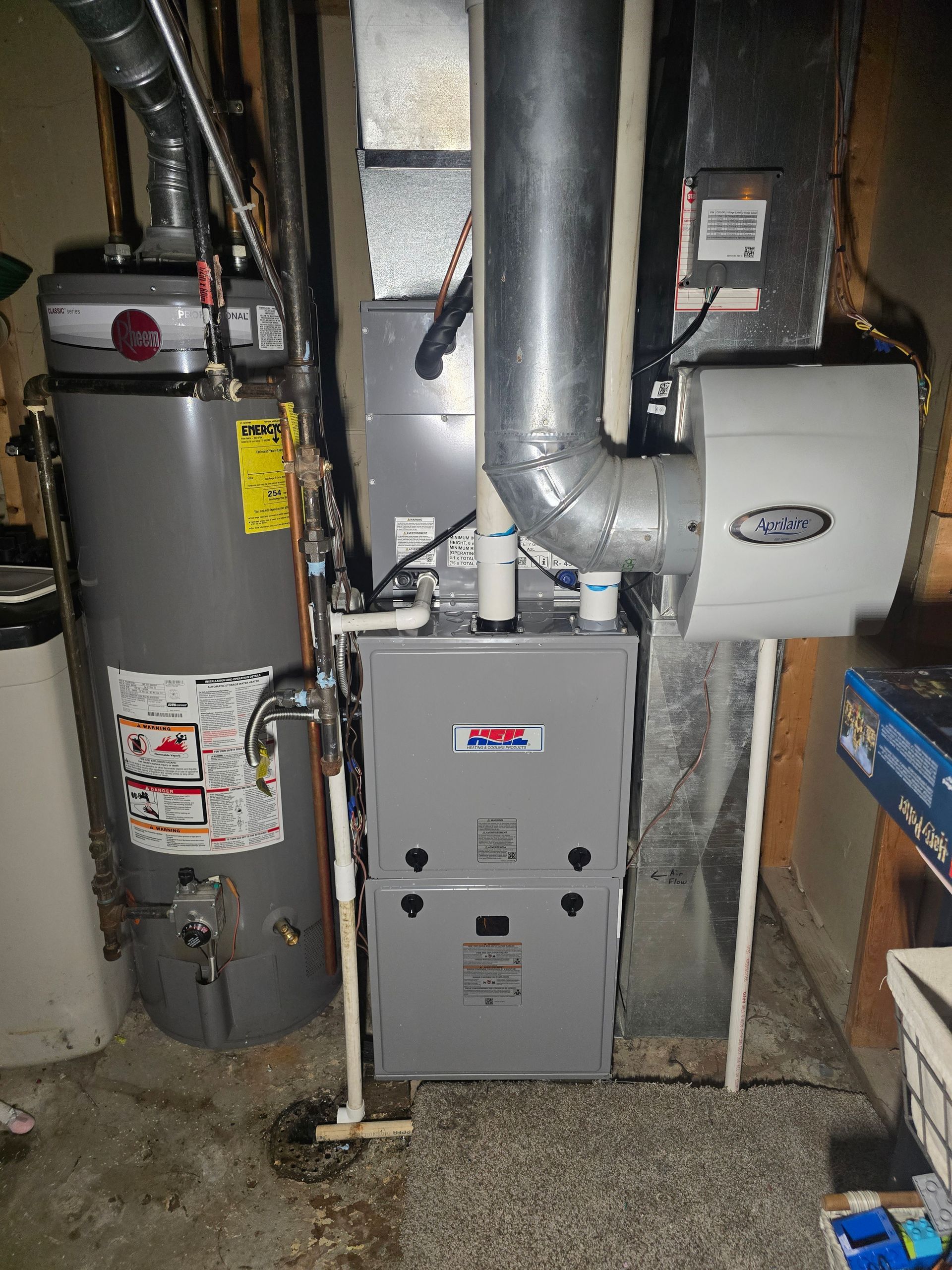 A gray water heater stands next to a furnace and a white humidifier attached to metal ductwork in a basement setting.