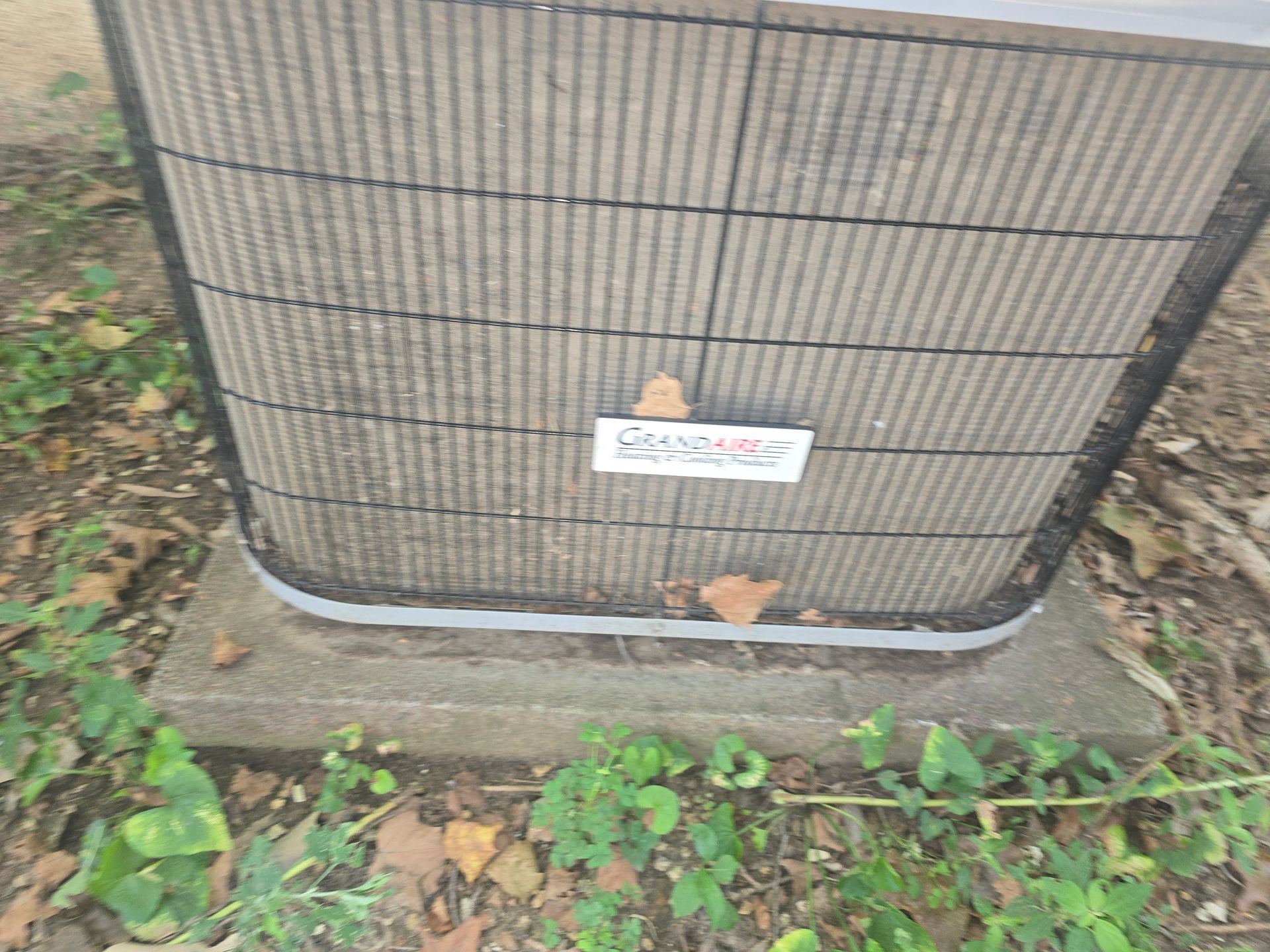 An outdoor air conditioning unit sits on a concrete pad surrounded by dirt and green ground cover.