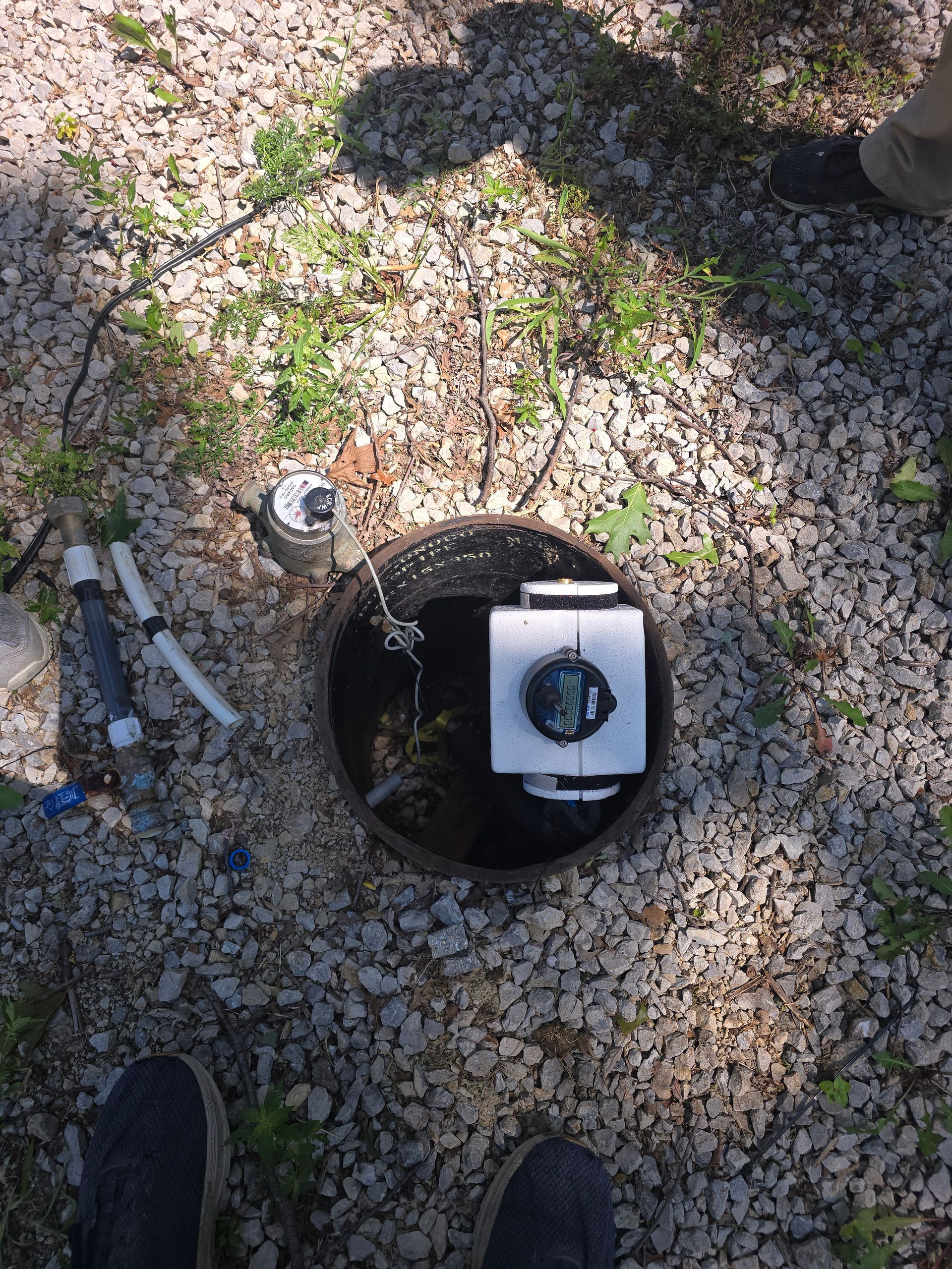 A water meter box set in light-colored gravel ground, showing a white meter assembly and exposed utility pipes.
