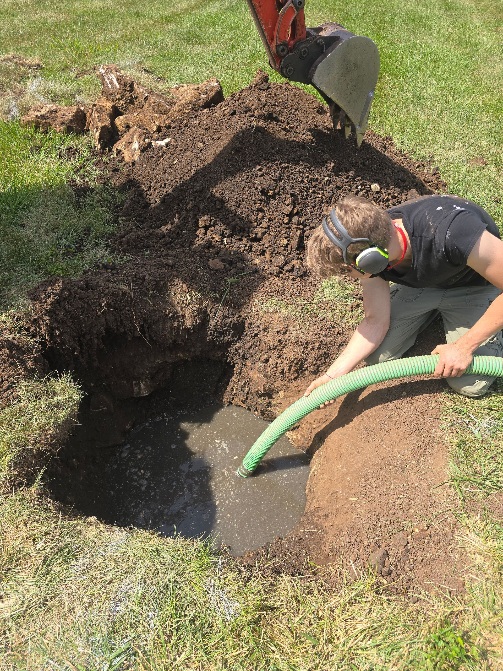 A person using a hose to pump water from an excavated hole in the ground while an excavator bucket hovers nearby.