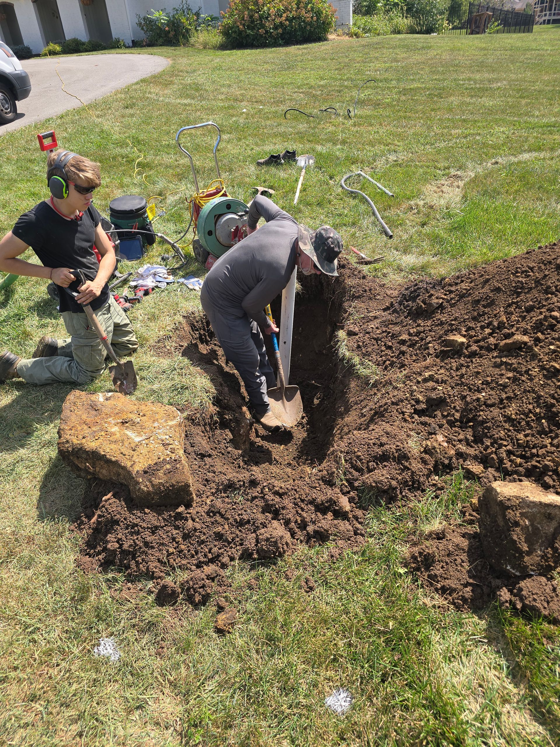 Two people in a grassy yard, one standing with headphones and the other kneeling and digging in a hole.