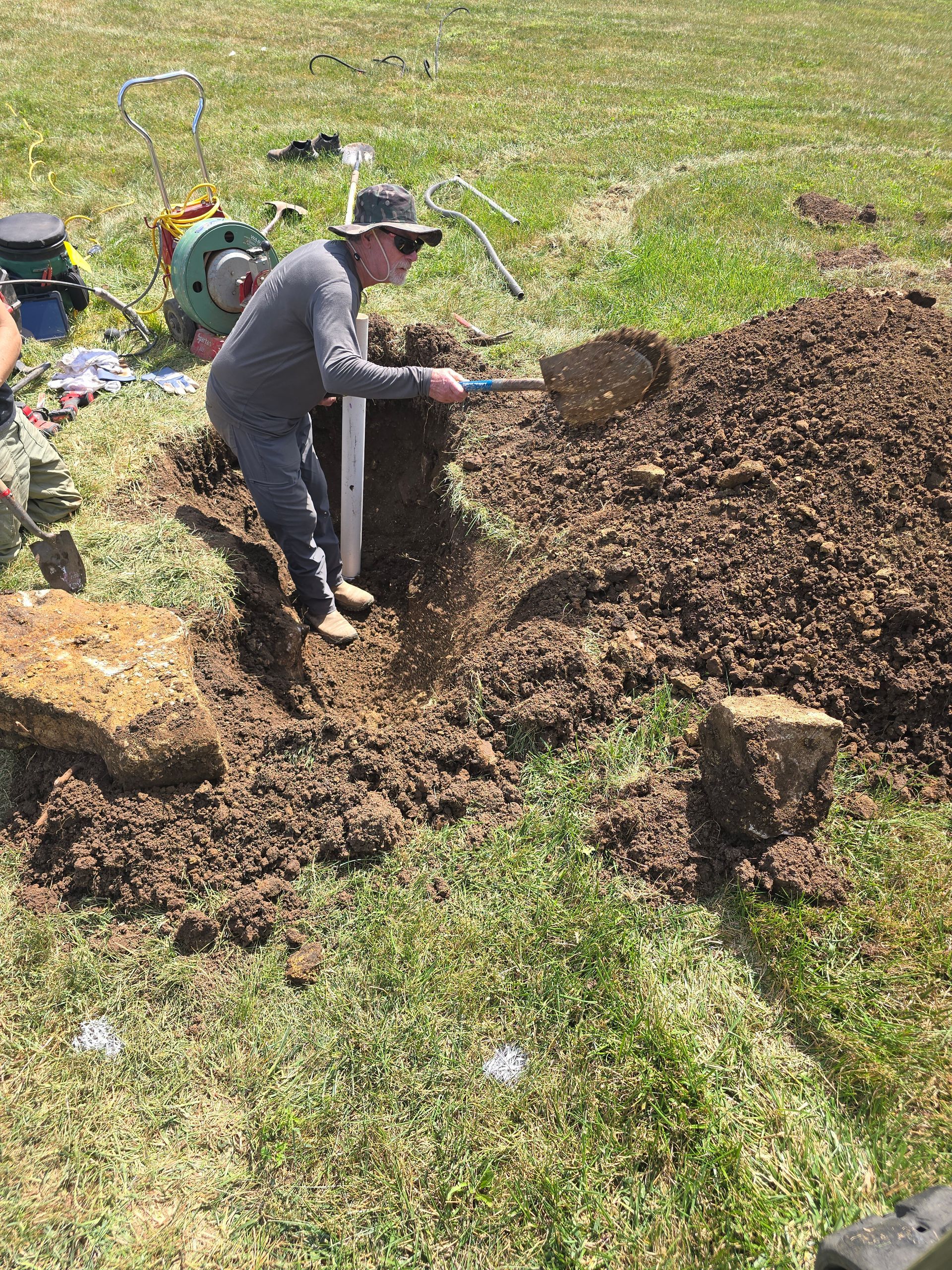 A person wearing a hat and grey clothing uses a shovel to dig in a grassy field next to an exposed white pipe.