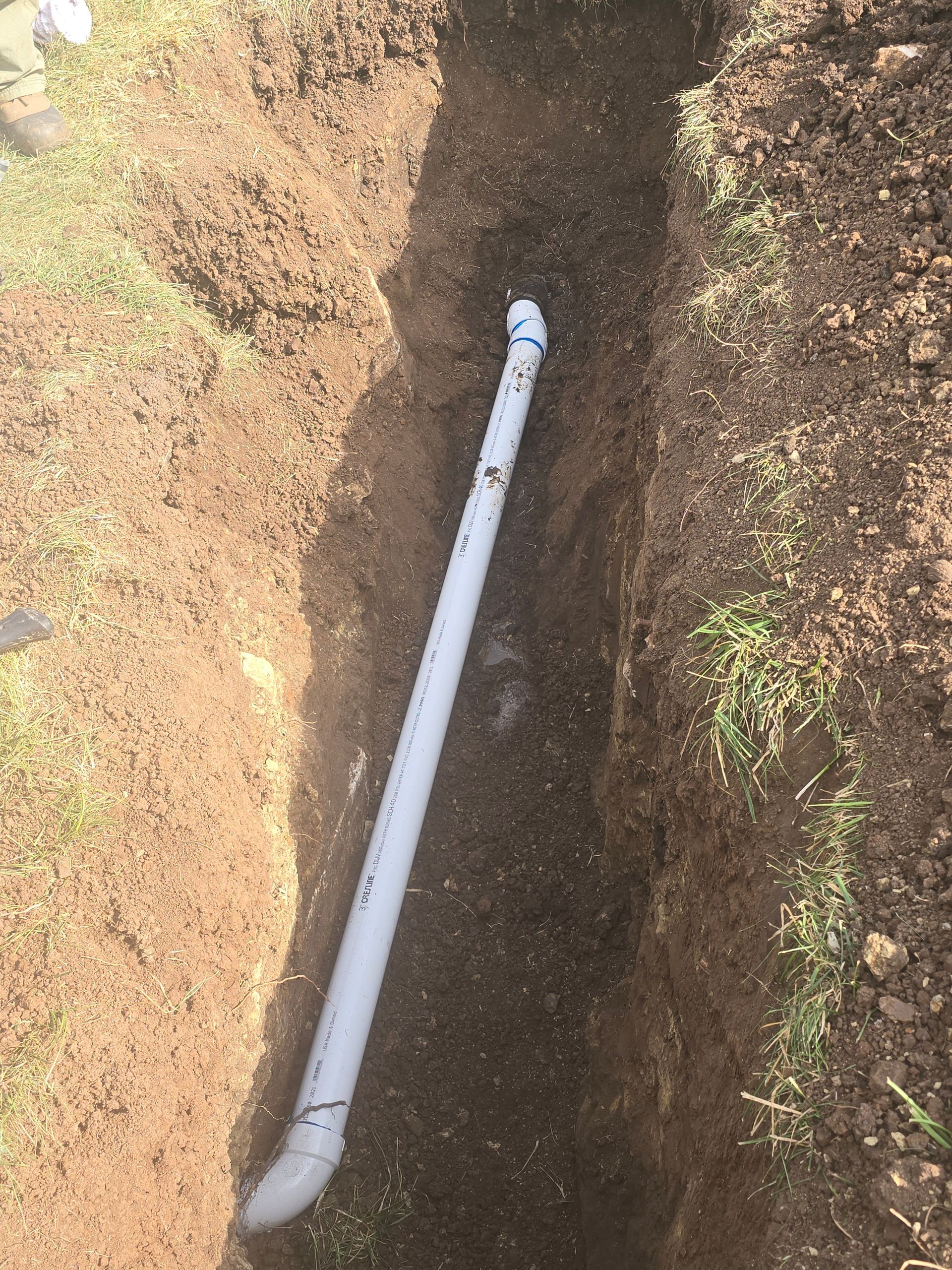 A white PVC pipe is laid inside a narrow, freshly dug dirt trench in an outdoor setting.