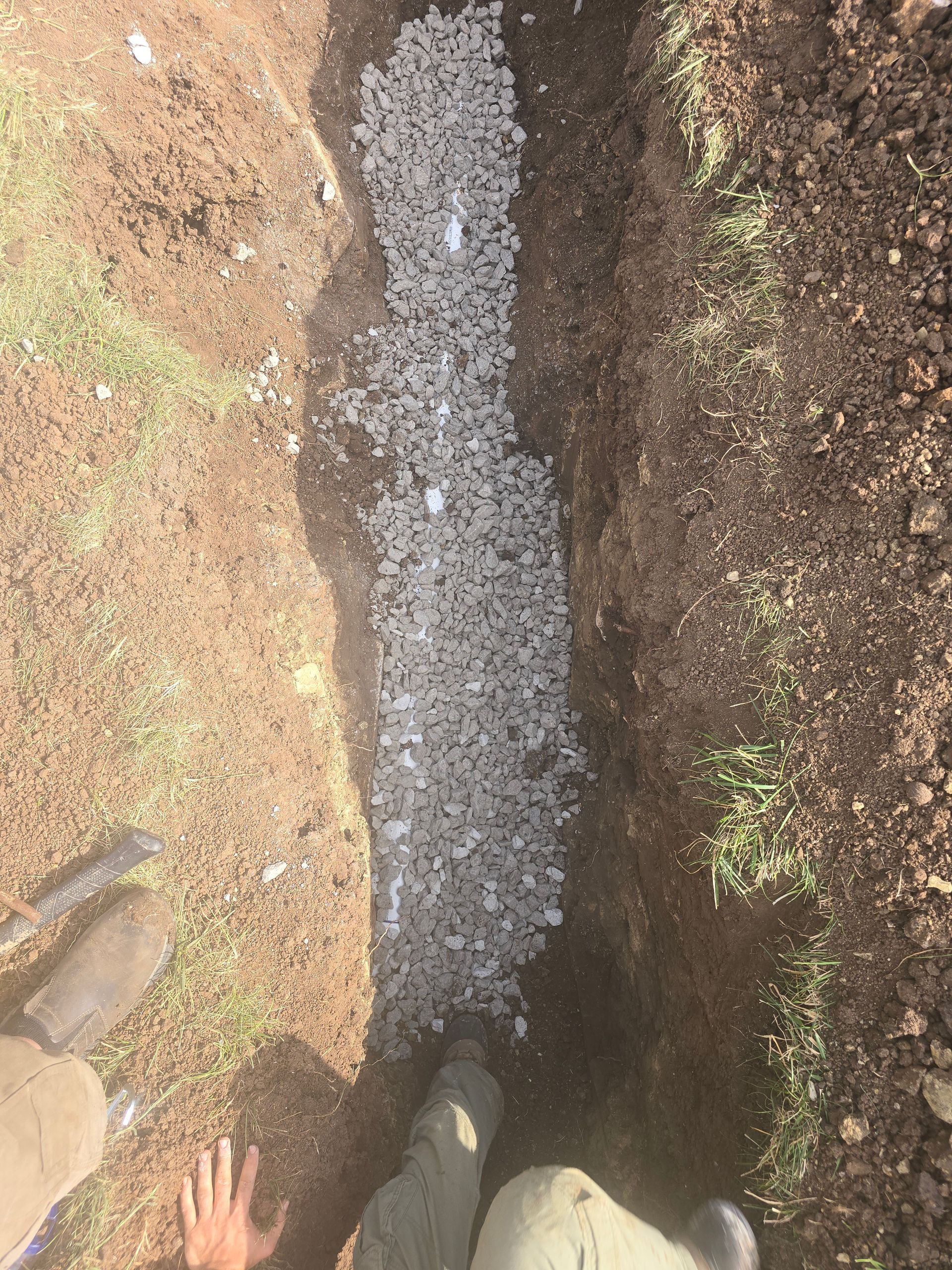 A top-down view of a trench filled with gray gravel, with a person's legs and hand visible at the bottom edge.