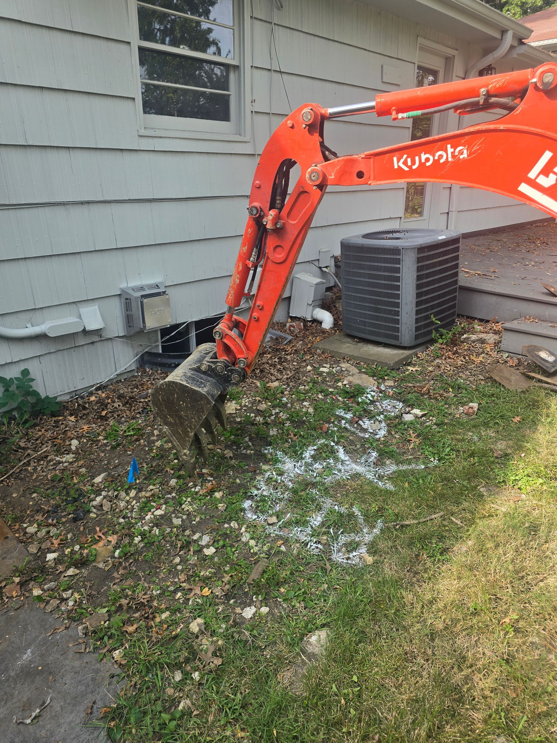 An orange Kubota excavator arm and bucket positioned over a marked patch of grass next to a house and air conditioner.