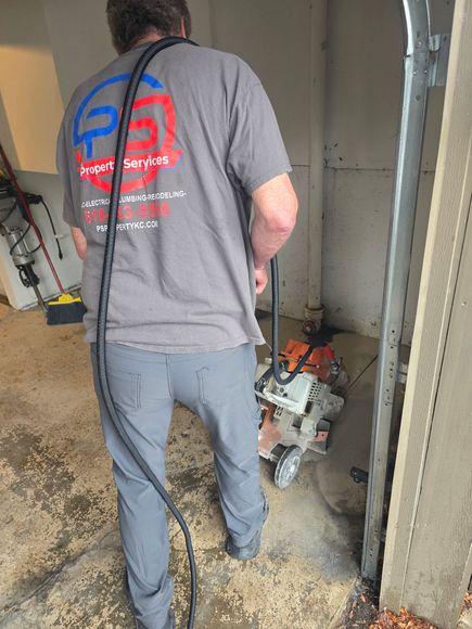 A person in a gray t-shirt operates a heavy-duty floor saw in a garage, holding a long black cable over their shoulder.