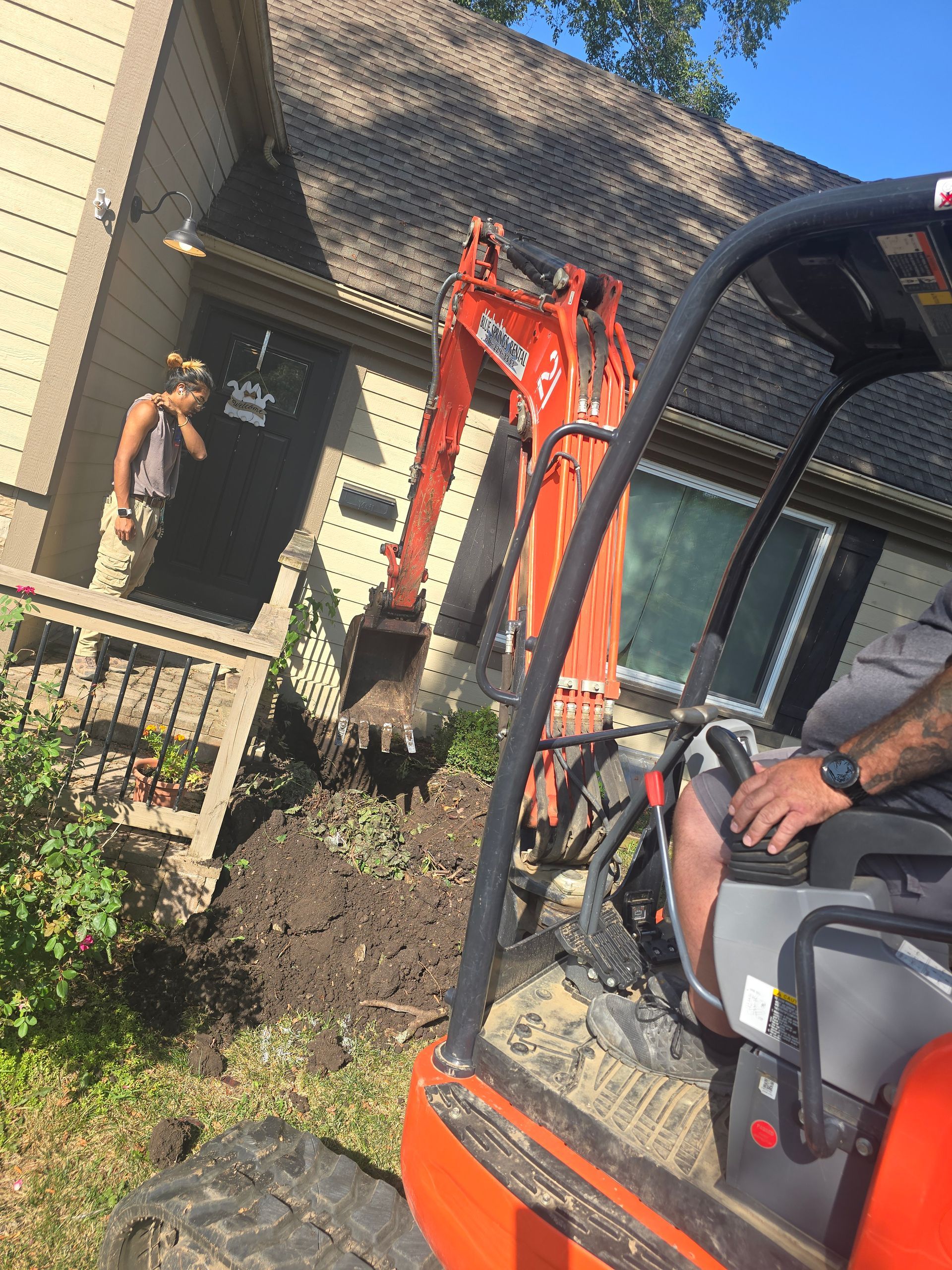 An orange excavator works in the front yard of a house while a person stands near the front entrance.
