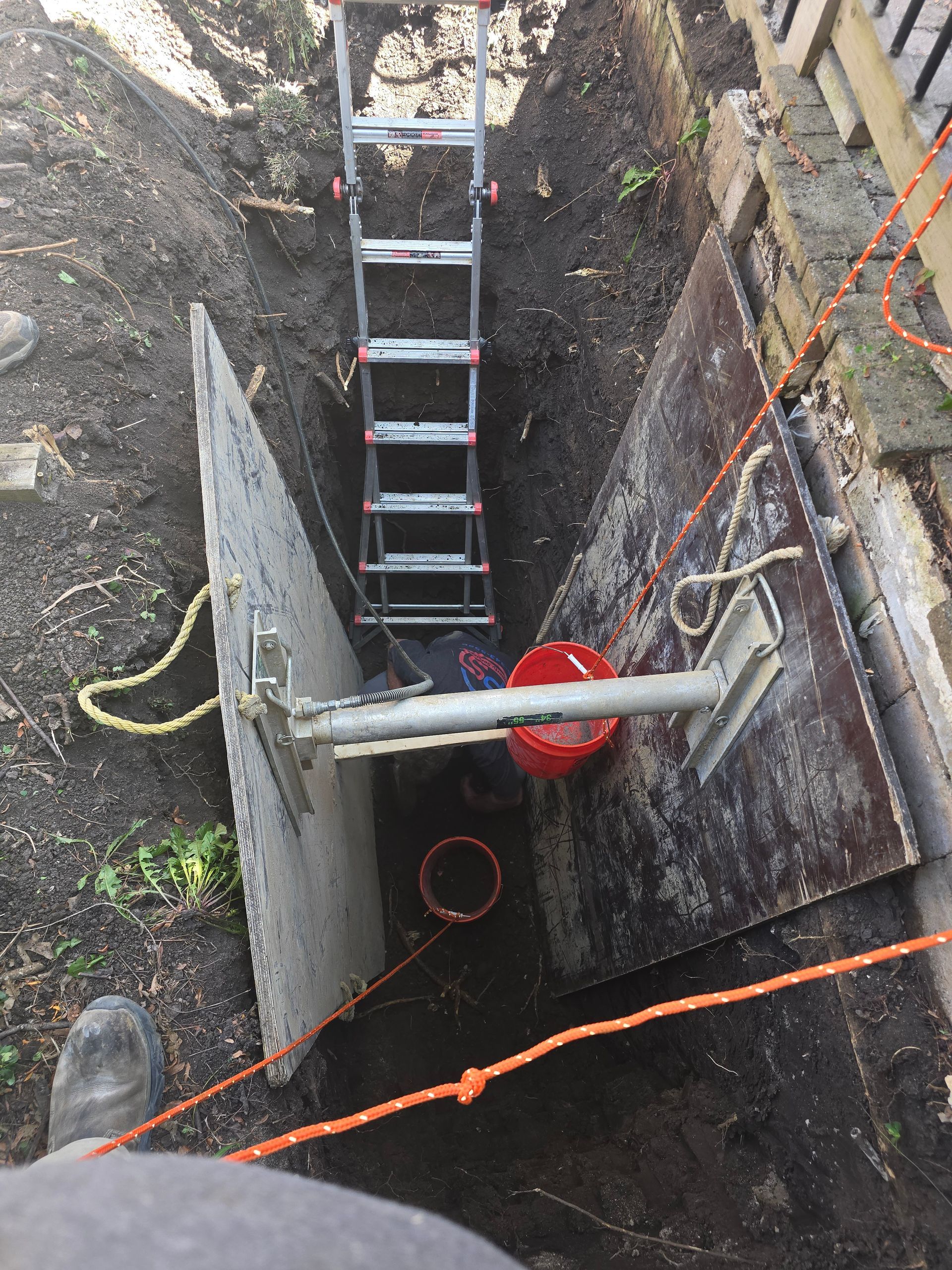 A person works inside a deep, shored excavation trench fitted with a metal ladder and a hydraulic brace.