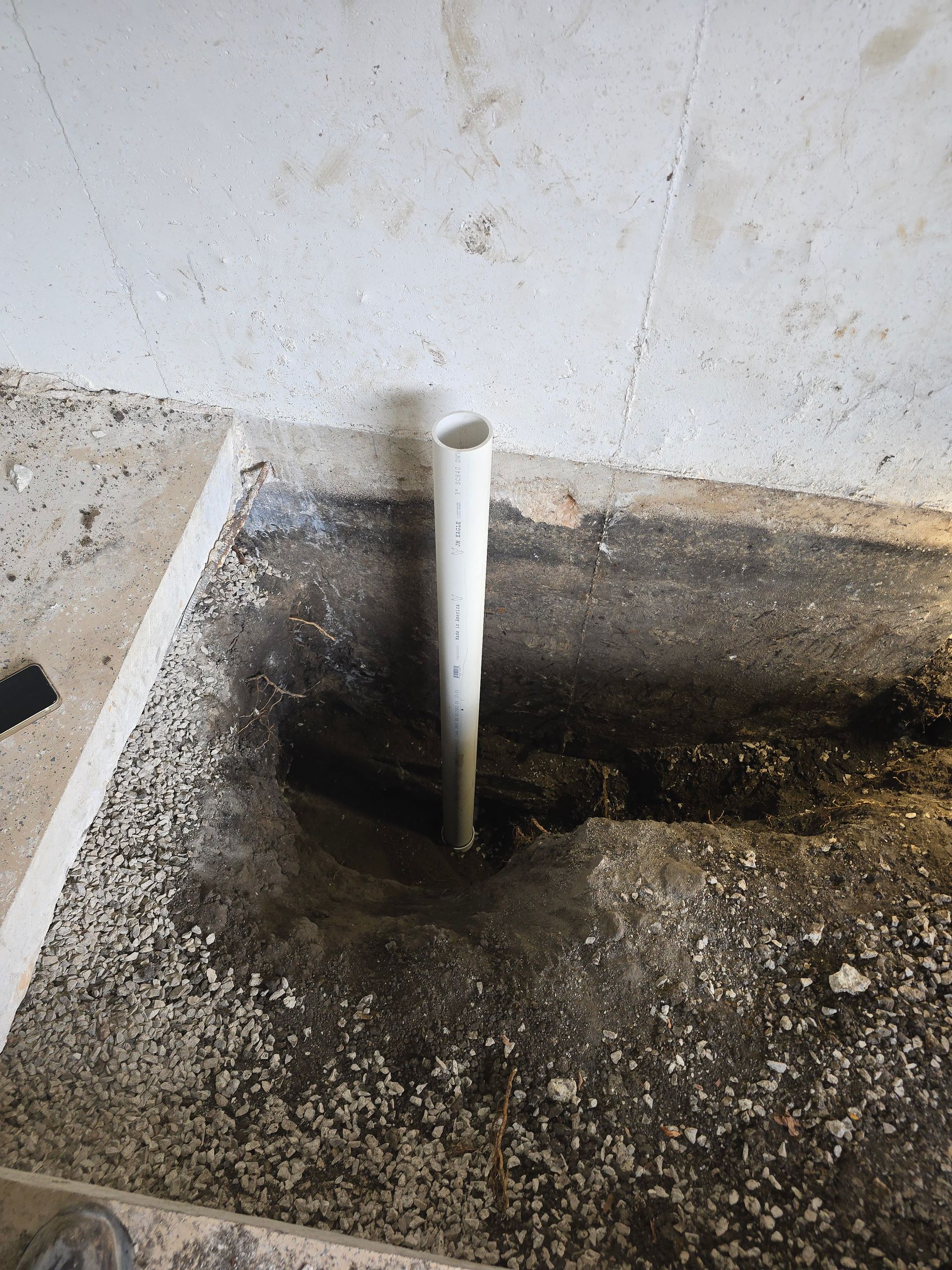 A white plastic pipe stands upright in a small, rectangular hole cut into a concrete floor, revealing dirt underneath.